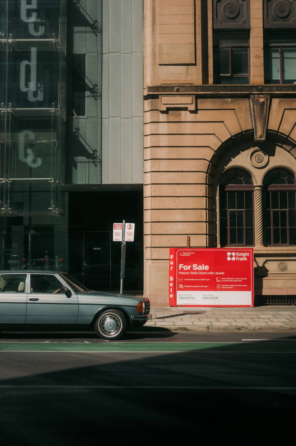 A pale car beside a red sign and a stone building.