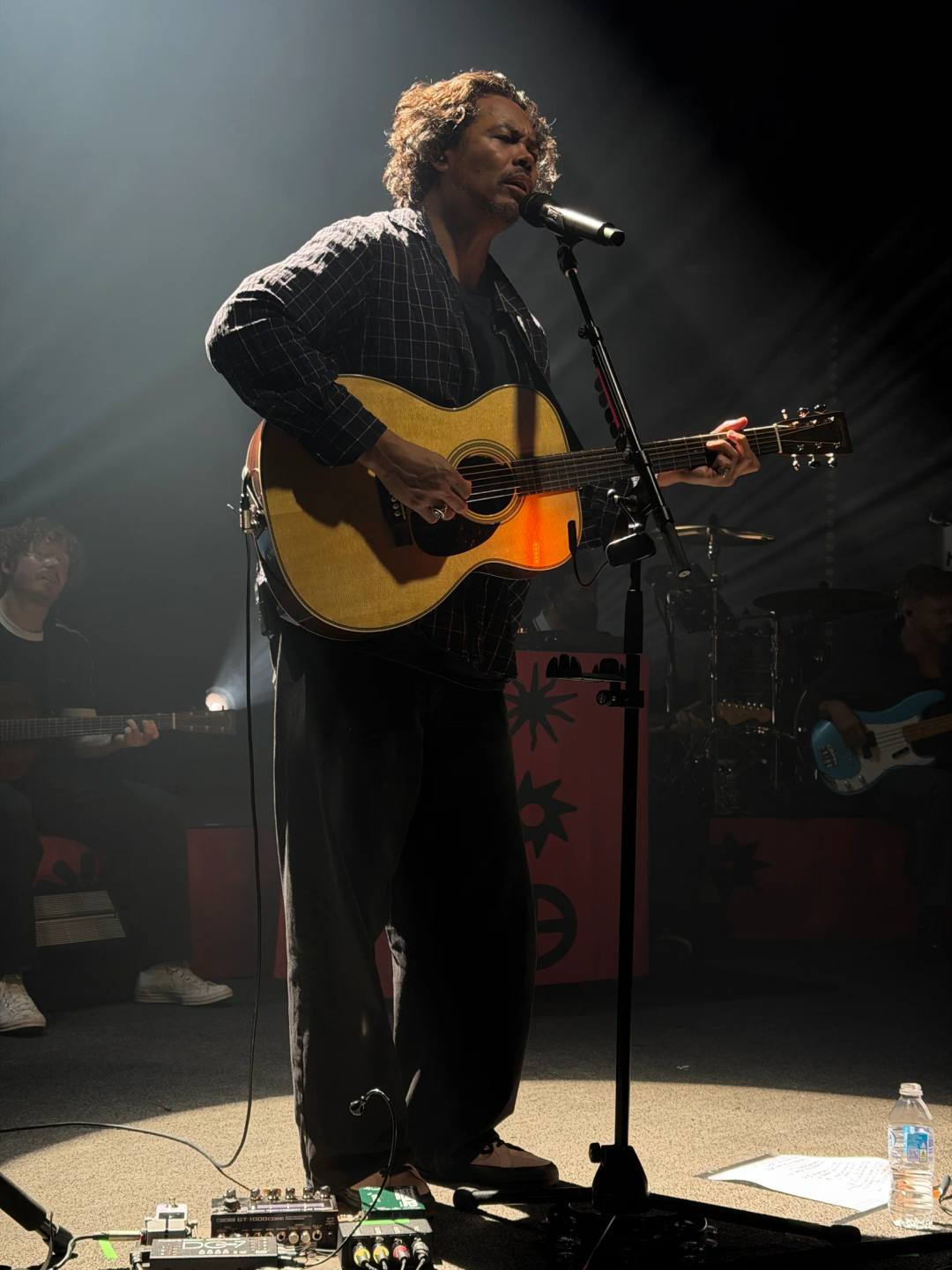 A full-length view of a singer playing an acoustic guitar at a microphone, with band members in the shadows behind.