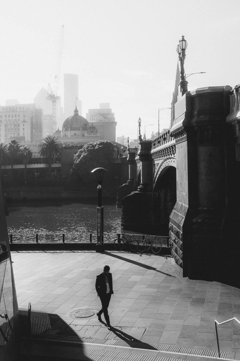 A pedestrian and long shadows beside a bridge, in black and white.