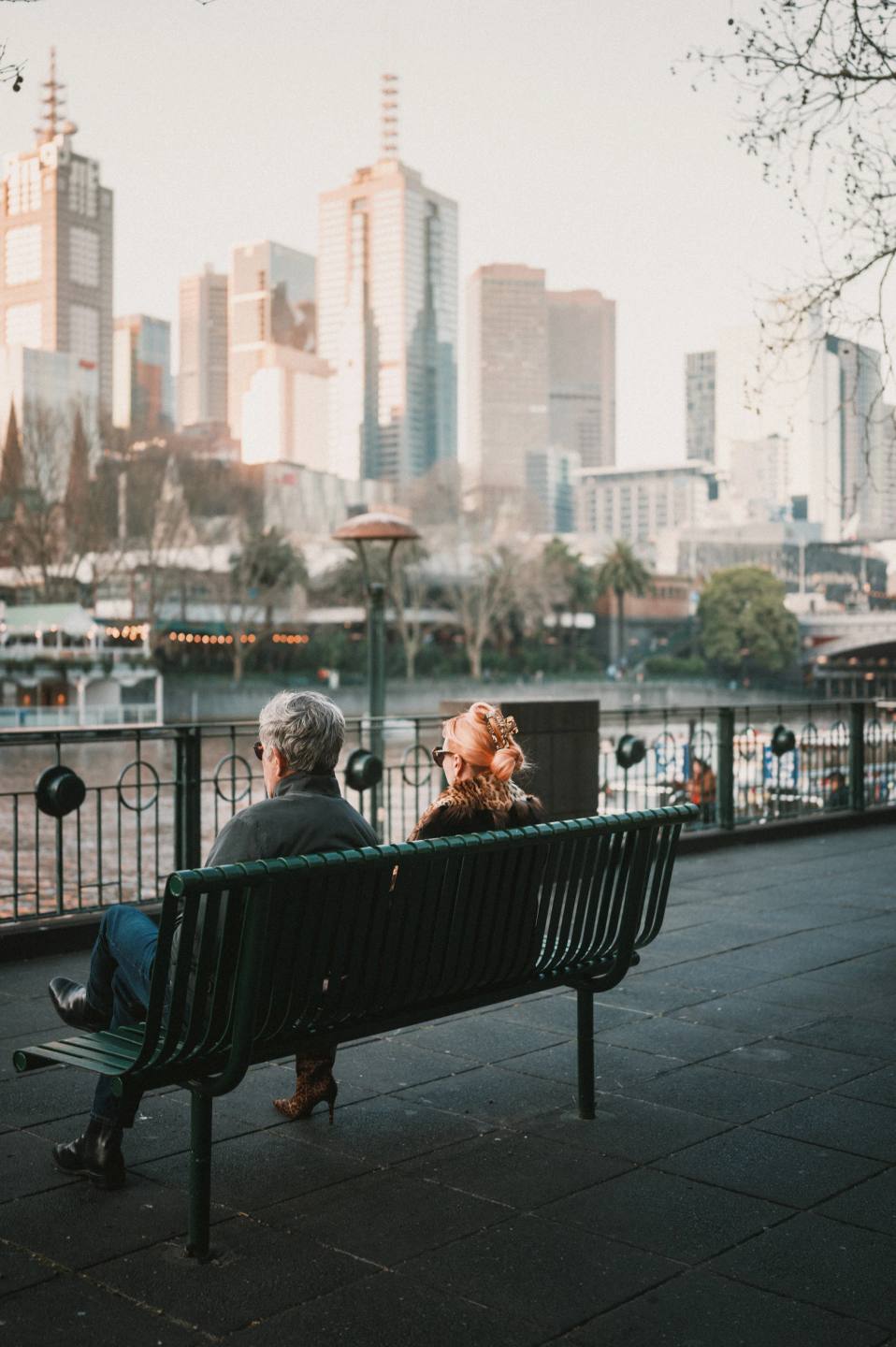 Two people sitting on a riverside bench facing the city.