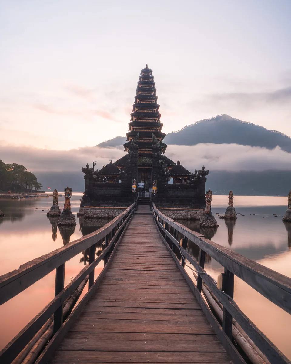 A wooden walkway leading to a lakeside temple at dawn.