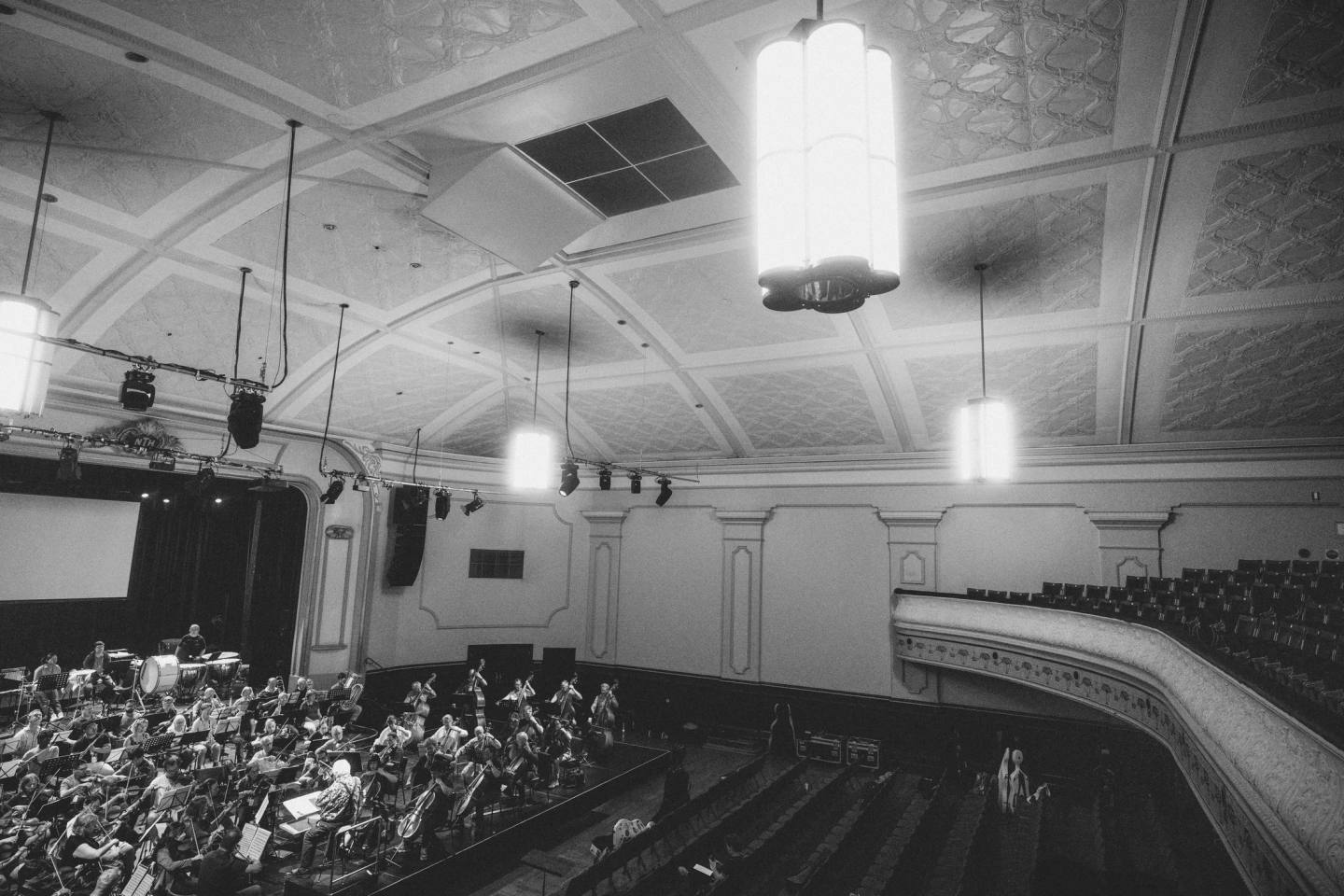A wide view of the orchestra beneath the concert hall ceiling and pendant lights, in black and white. Australian Doctors Orchestra 2026, Adelaide.