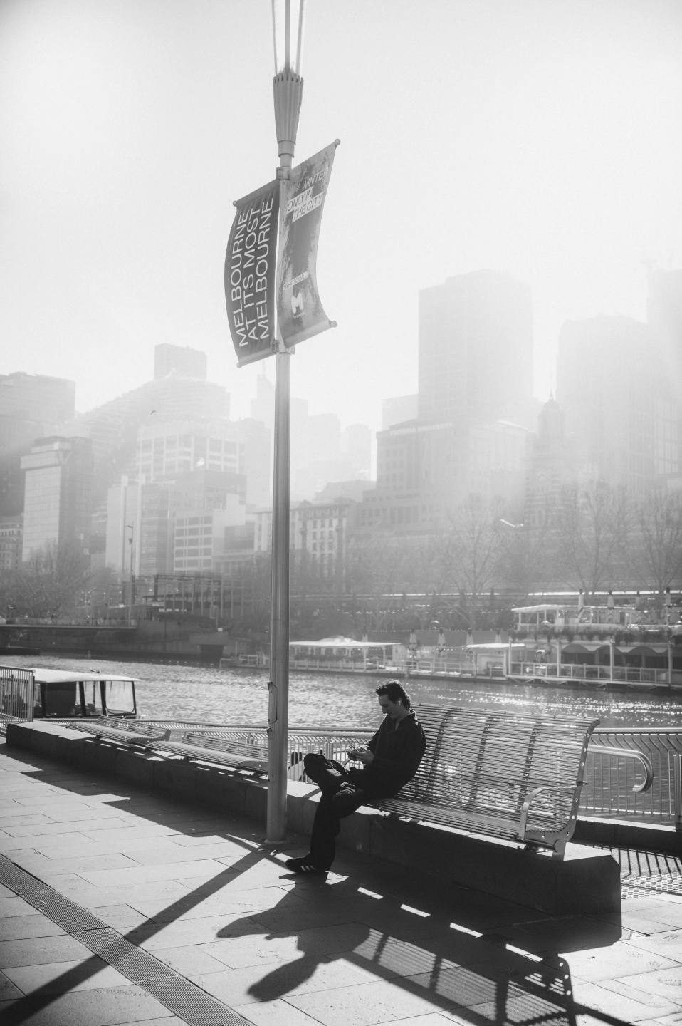 A person on a riverside bench against a hazy city skyline.
