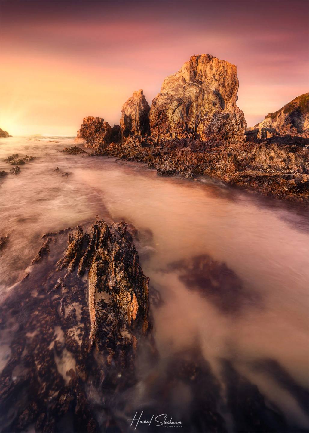 Jagged coastal rocks surrounded by soft, flowing water beneath a pink sunset.