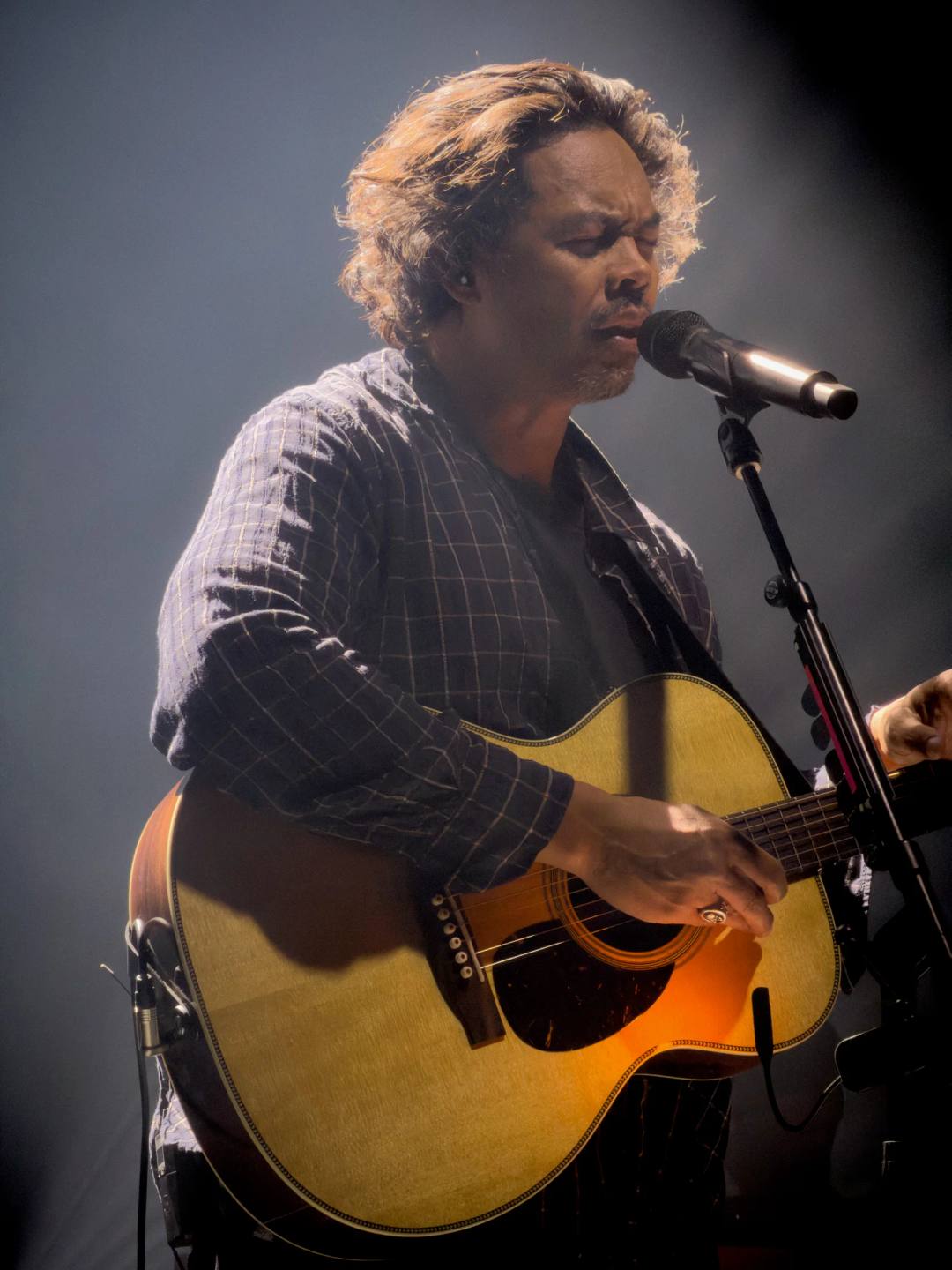 A singer playing an acoustic guitar at a microphone beneath a soft spotlight.