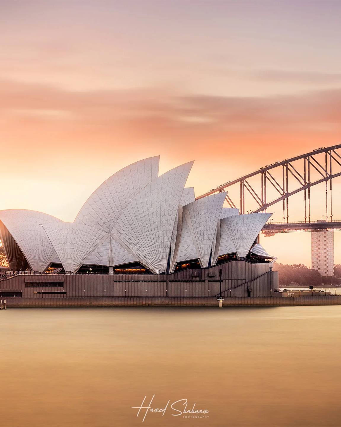 Sydney Opera House and Harbour Bridge reflected in calm water at sunset.