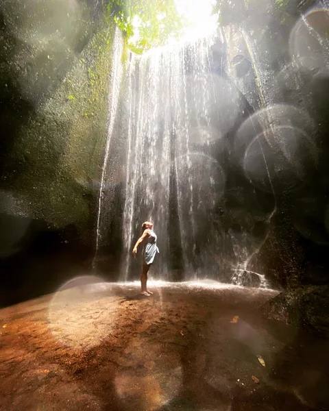 A person standing beneath a waterfall illuminated by sunlight in a rocky gorge.