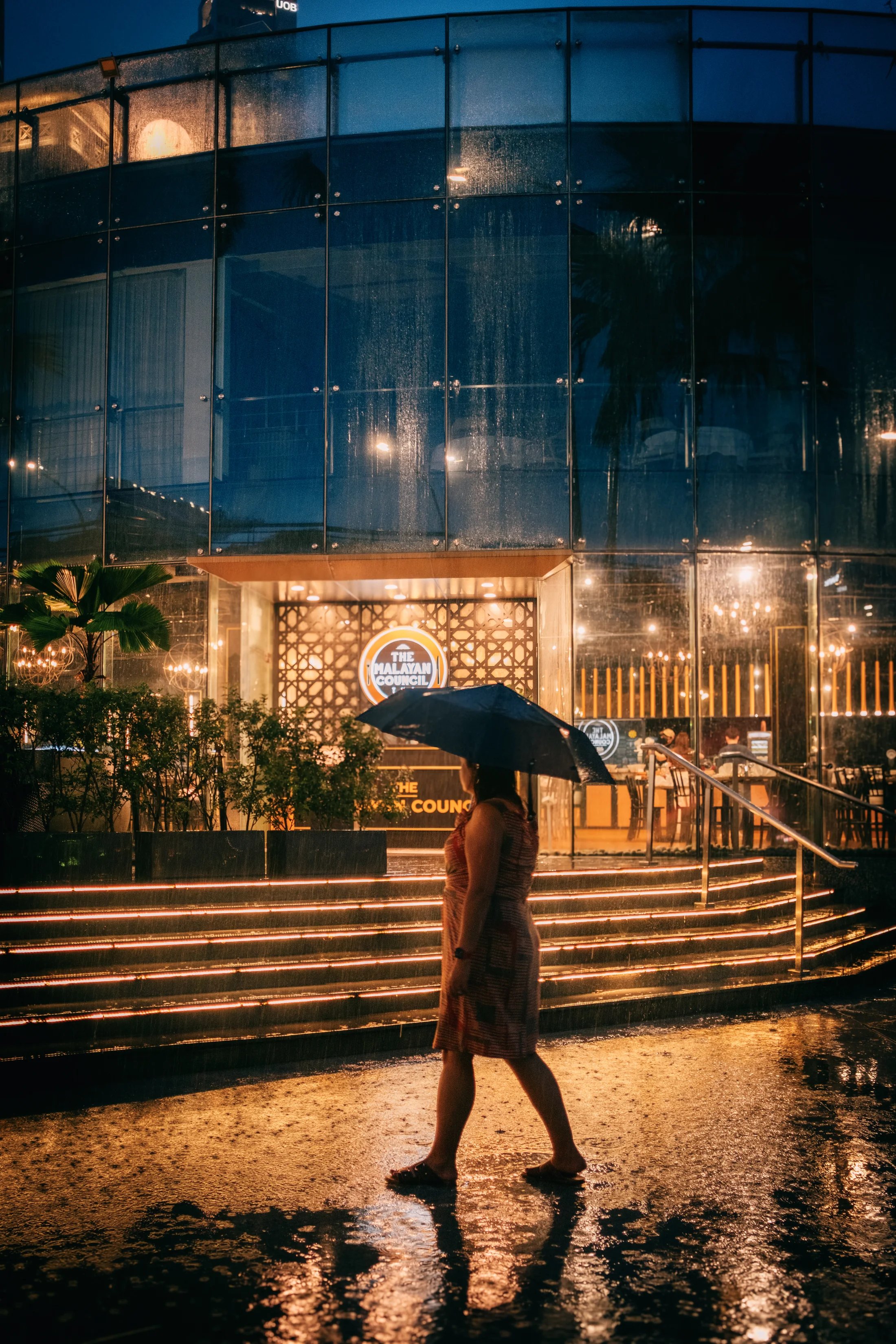 A person carrying an umbrella past warm lights reflected in the rain.