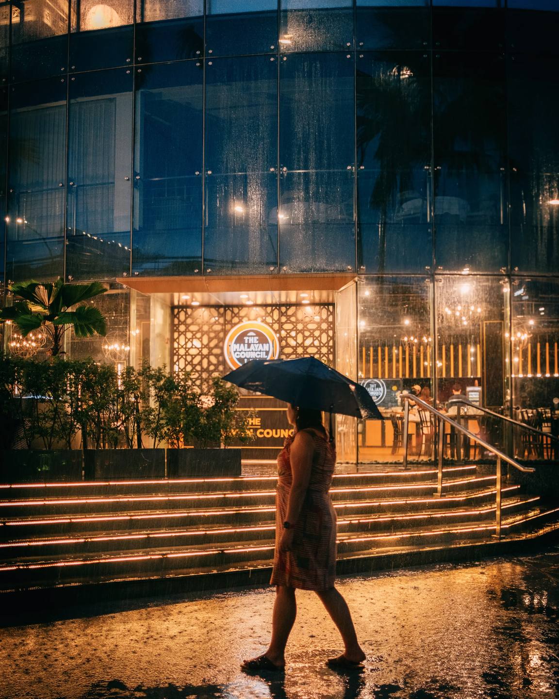A person carrying an umbrella past warm lights reflected in the rain.