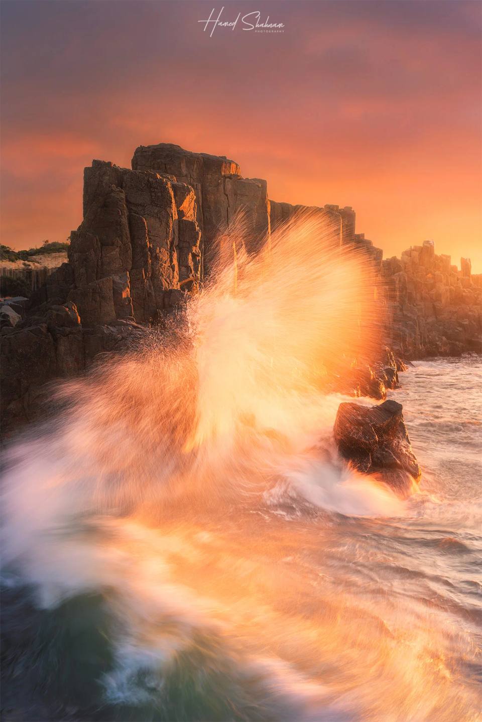 A wave breaking against dark rock columns in warm coastal light.