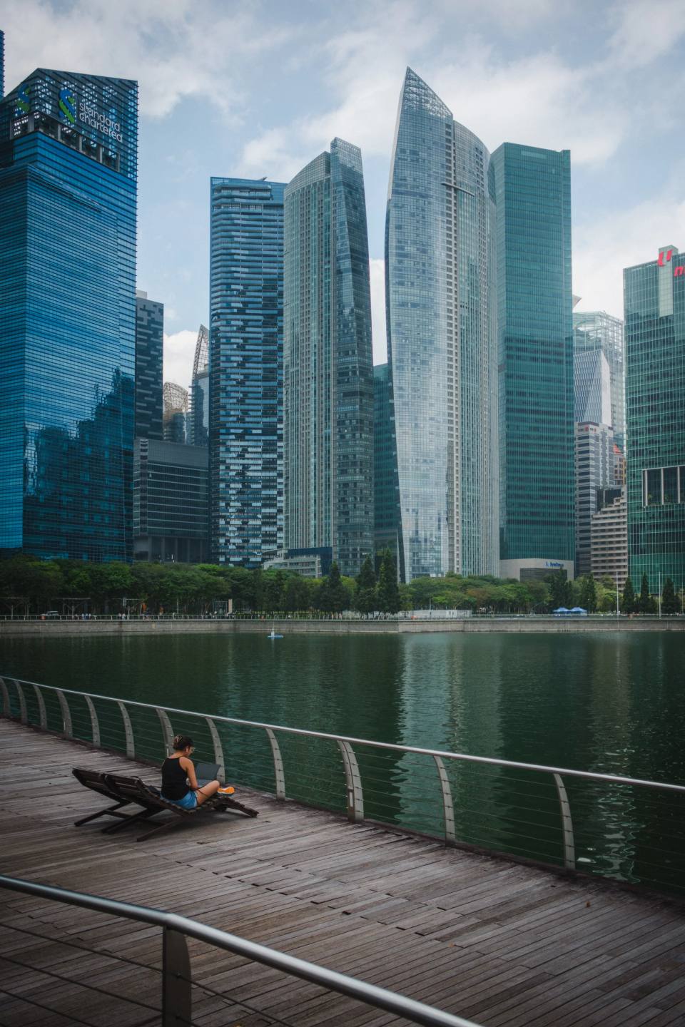 Skyscrapers reflected in water beside a riverside walkway.