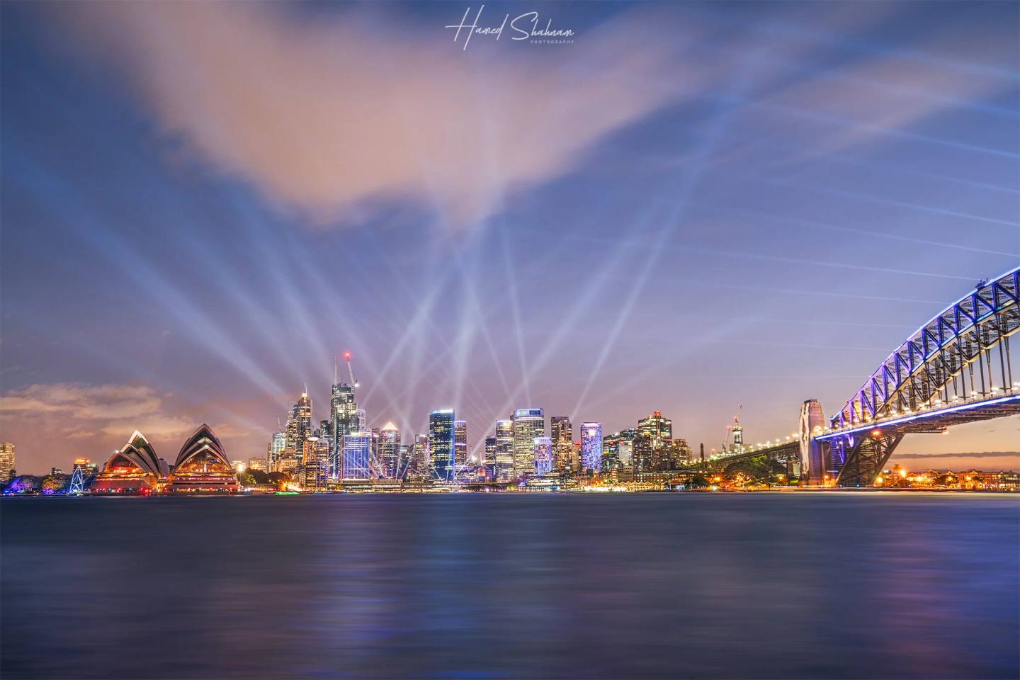 Beams of light above Sydney Harbour during Vivid, with the Opera House and Harbour Bridge.