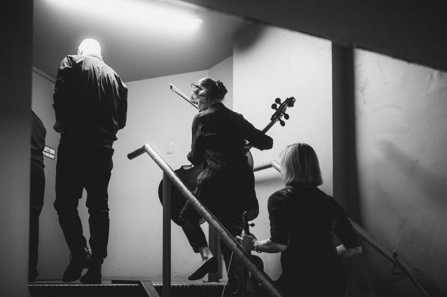 Musicians carrying string instruments up a backstage staircase, in black and white. Australian Doctors Orchestra 2026, Adelaide.