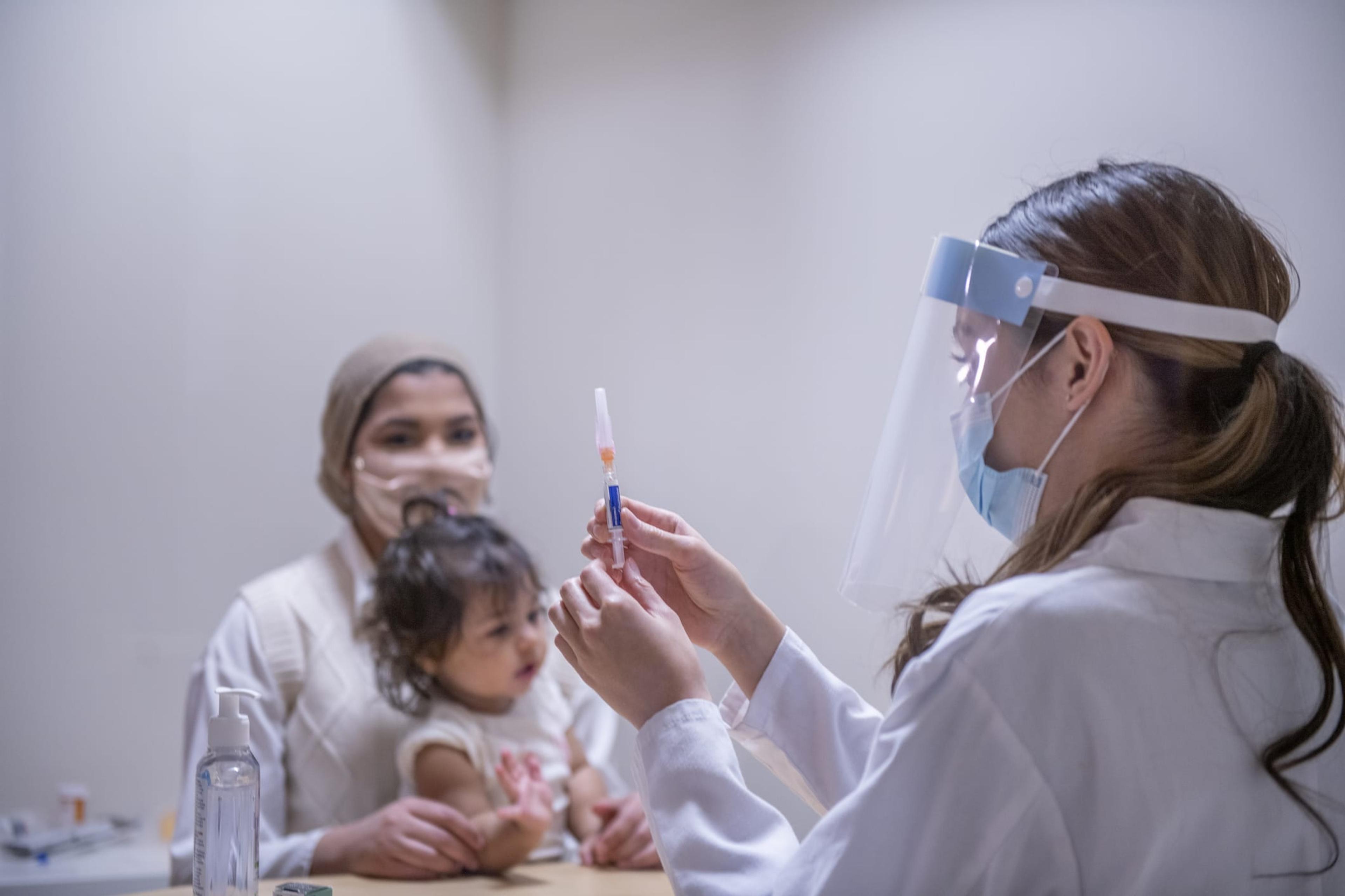 A Muslim mother wearing a hijab is with her young daughter at the doctor's office. The female doctor is preparing the medical syringe to administer the vaccine injection to her patients.