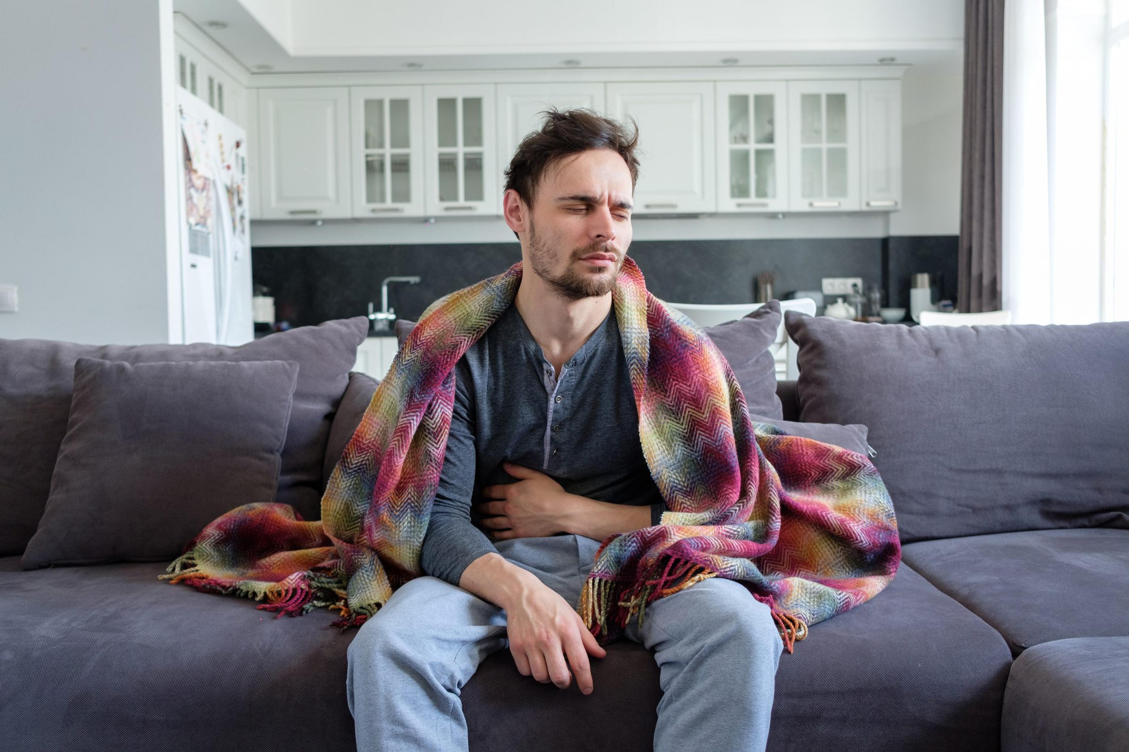Man sits on his couch clutching his stomach and wearing a blanket wondering if he has food poisoning or a stomach bug