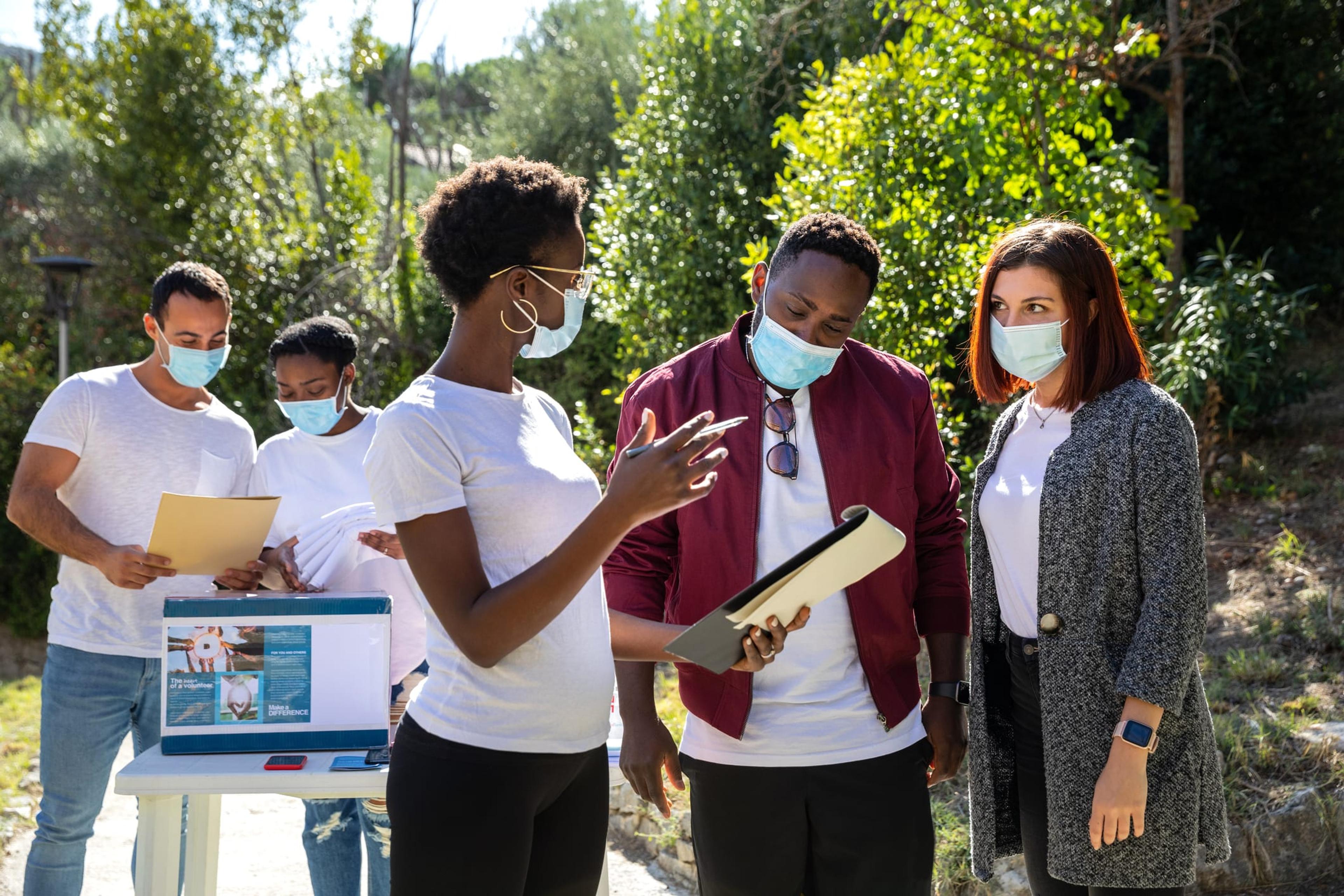 Diverse individuals in masks discuss something written on a notebook.