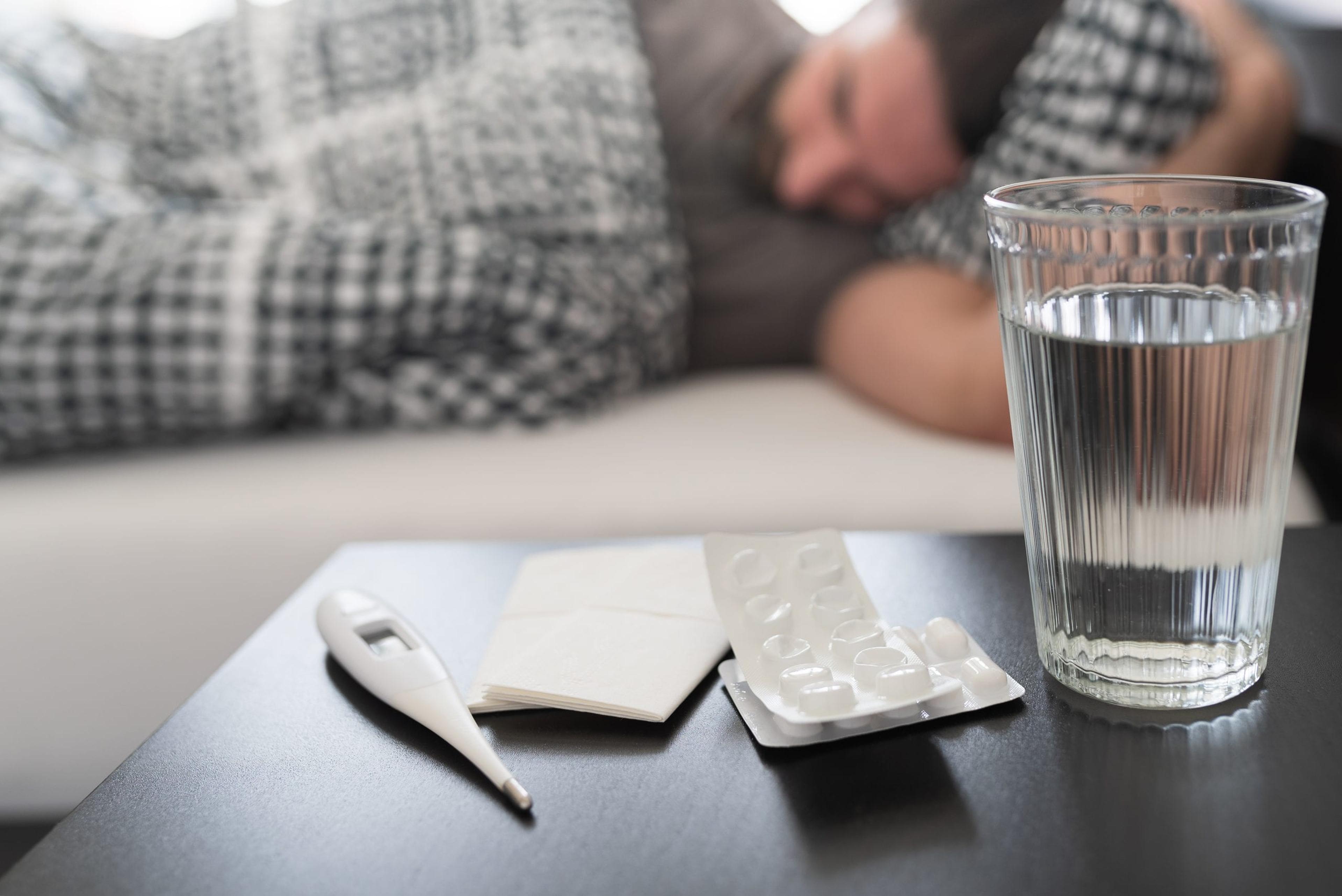 meds, medical thermometer and glass of water on nightstand against sick person suffering from flu or common cold sleeping in bed