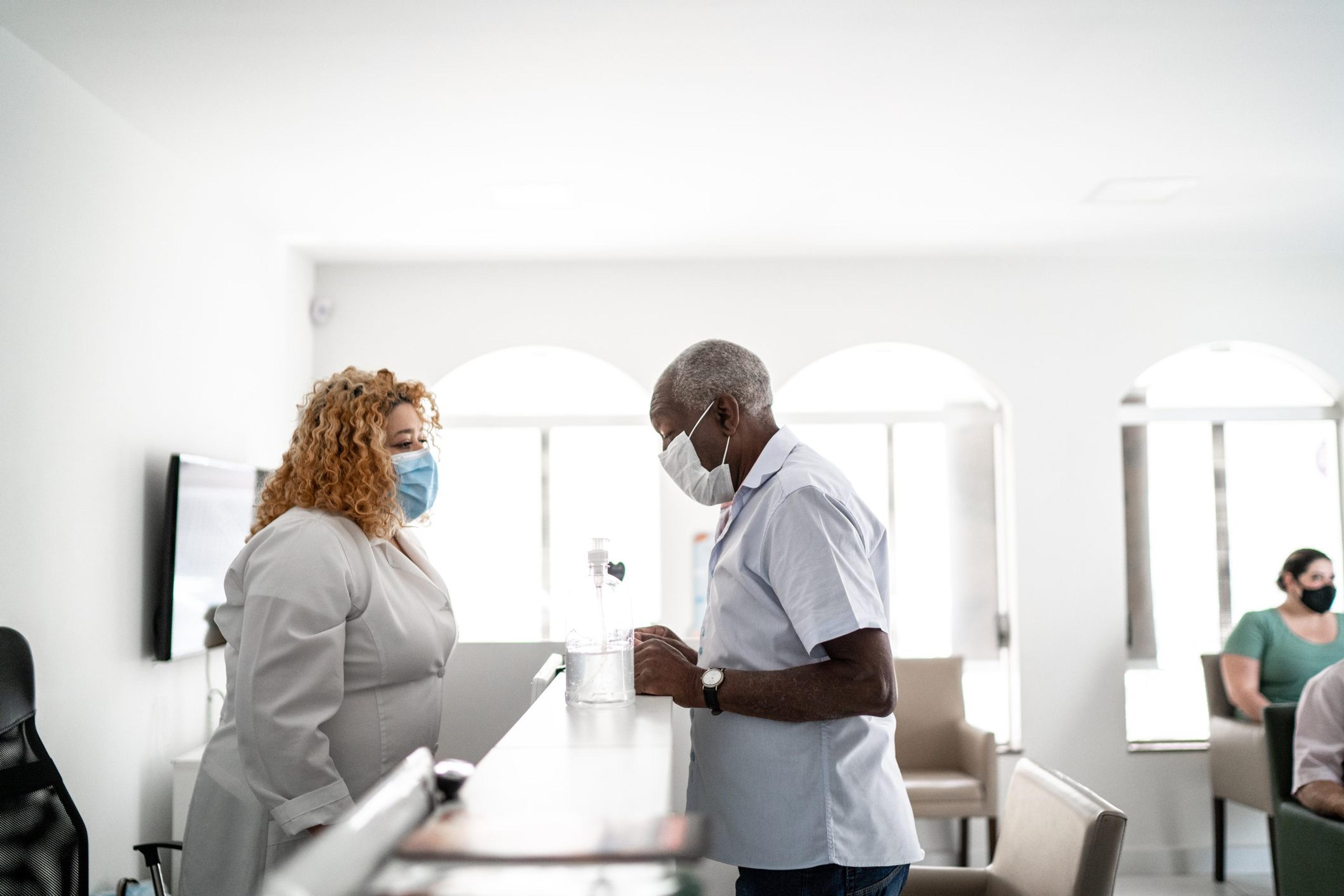 A medical clinic reception, receptionist talking to patient using face mask.