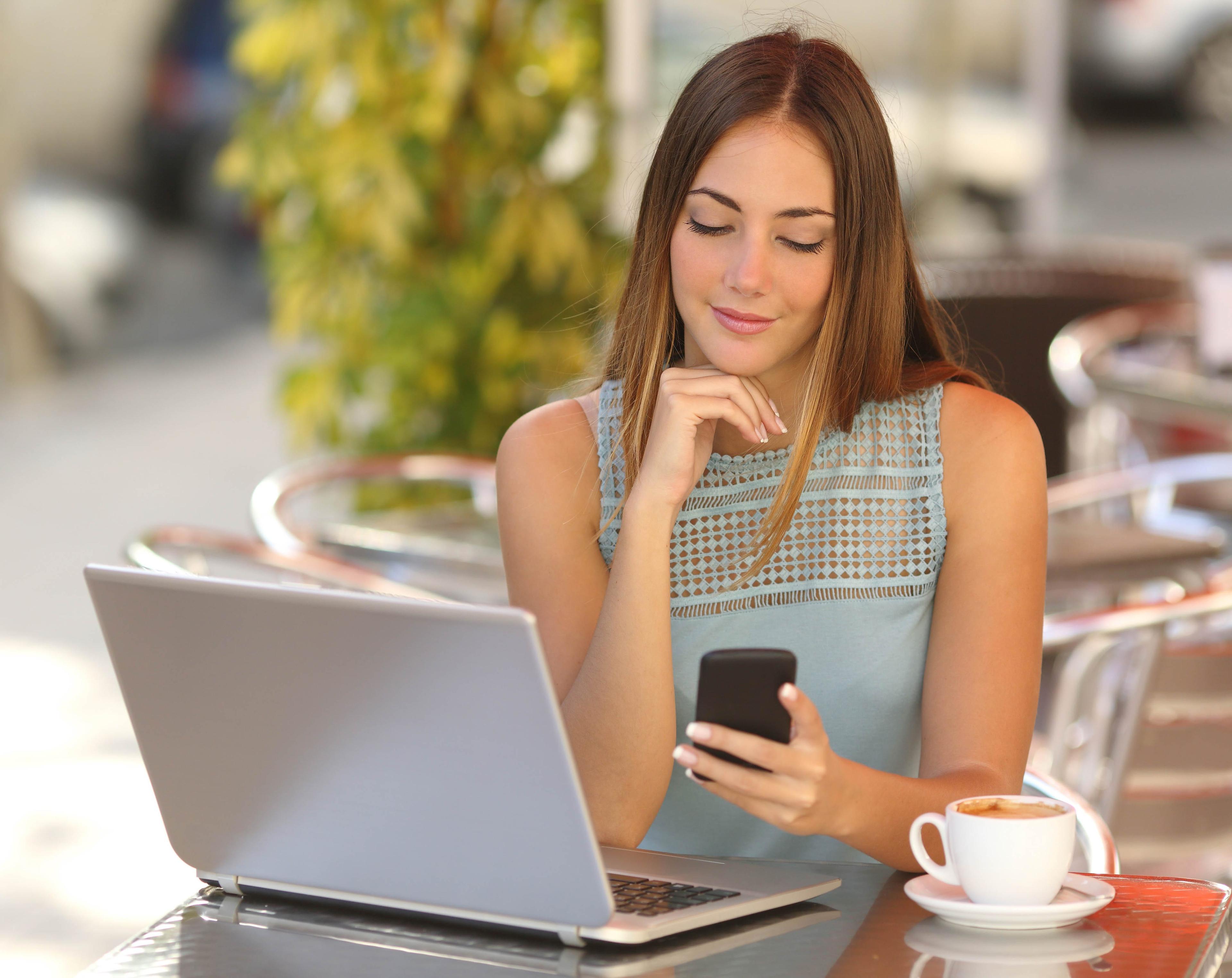 Self employed woman working with her phone and laptop in a restaurant terrace with a cup of coffee