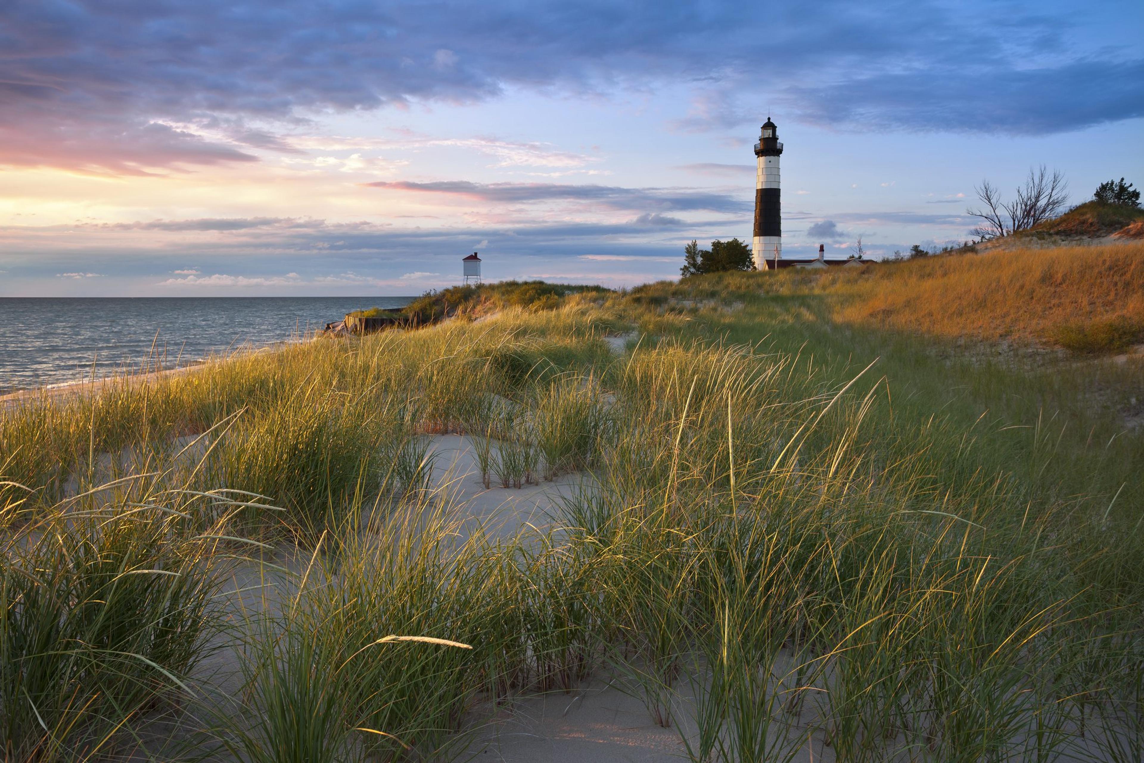 A lighthouse sits in the background of grassy beach as the sun sets.