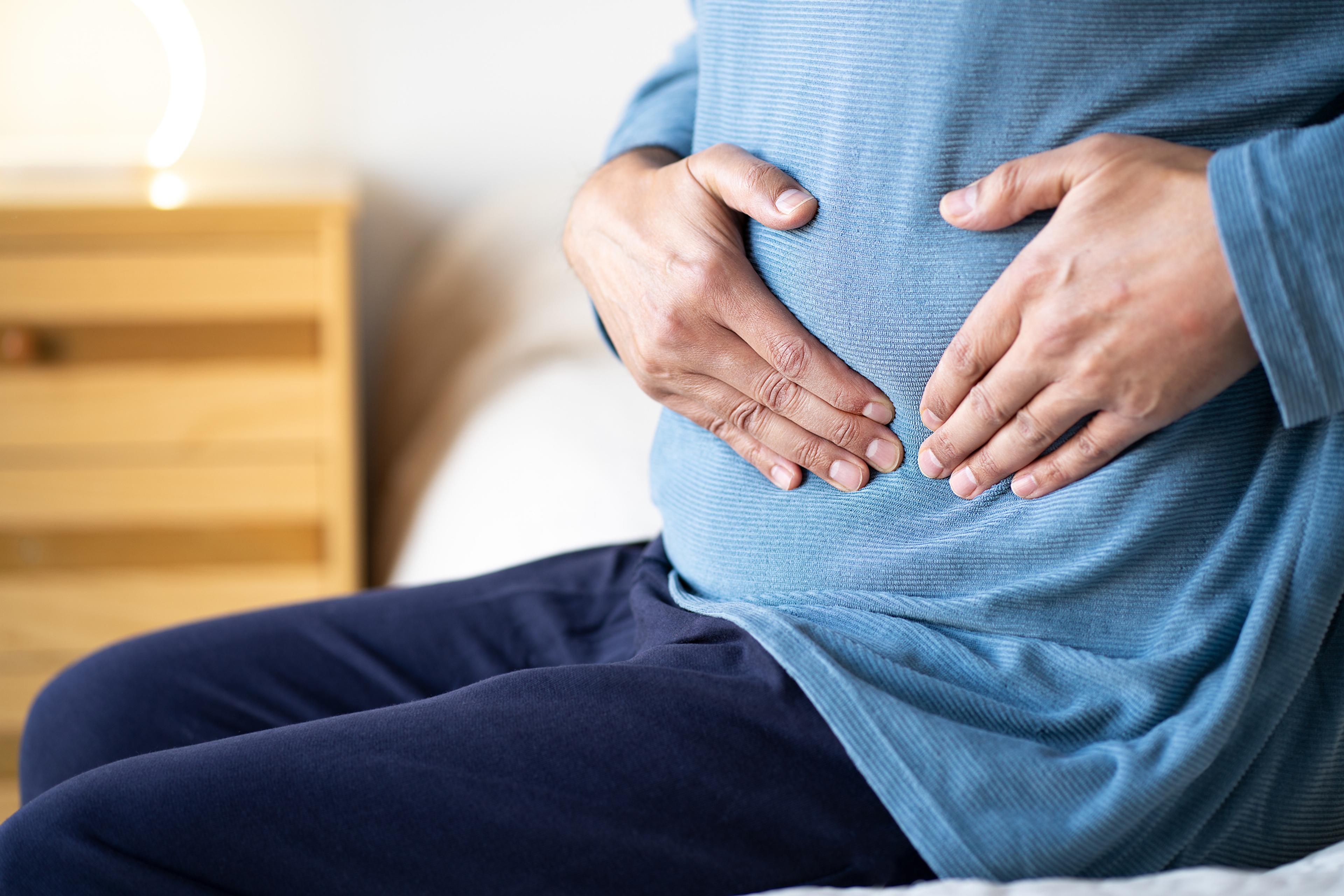 Man sitting alone on his bed and suffering from stomach cramps while home alone.Abdominal pain can be caused by digestive problems, infections, intestinal inflammation or more serious diseases.