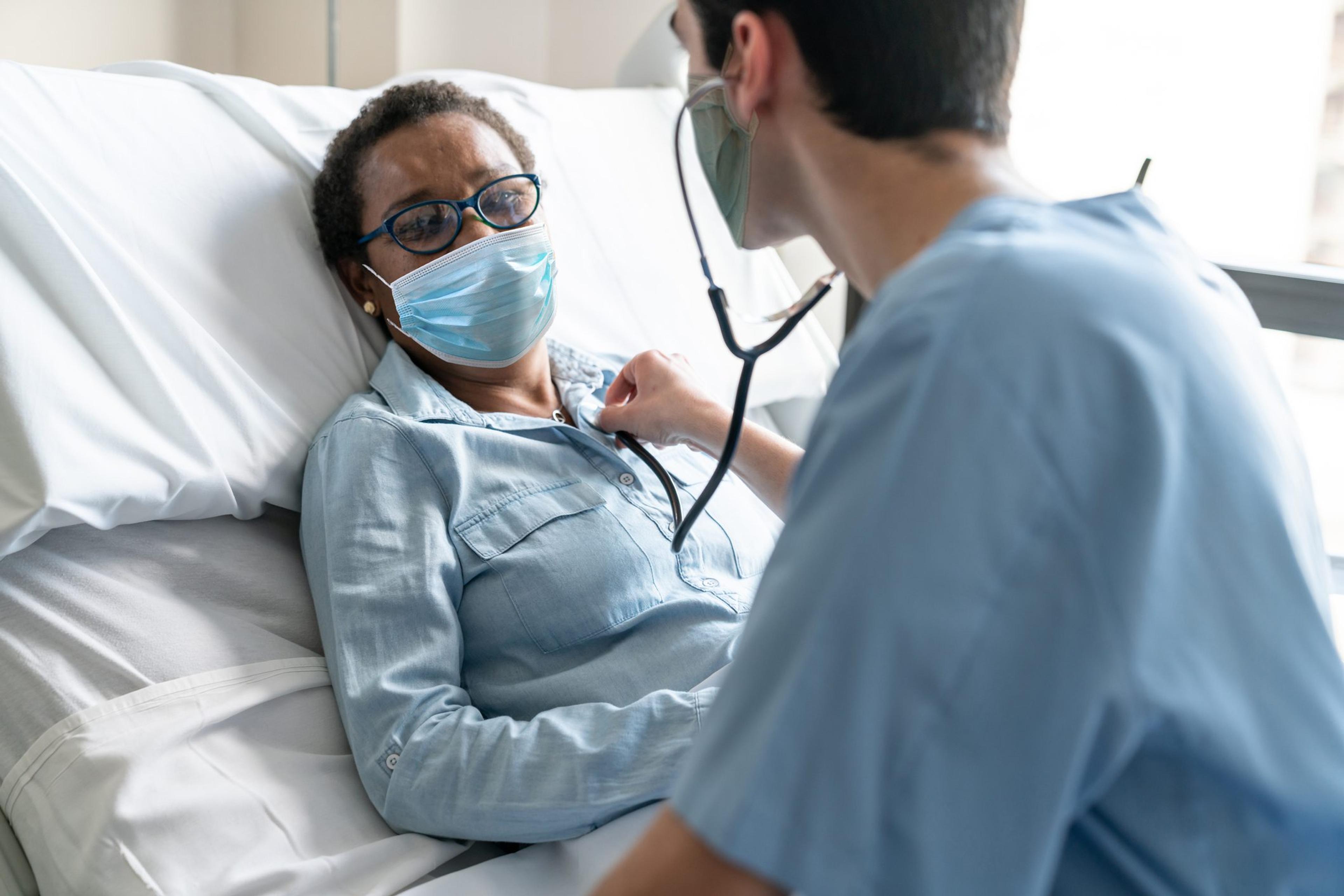 Latin american male nurse checking black female patient's heart with a stethoscope both wearing protective facemasks