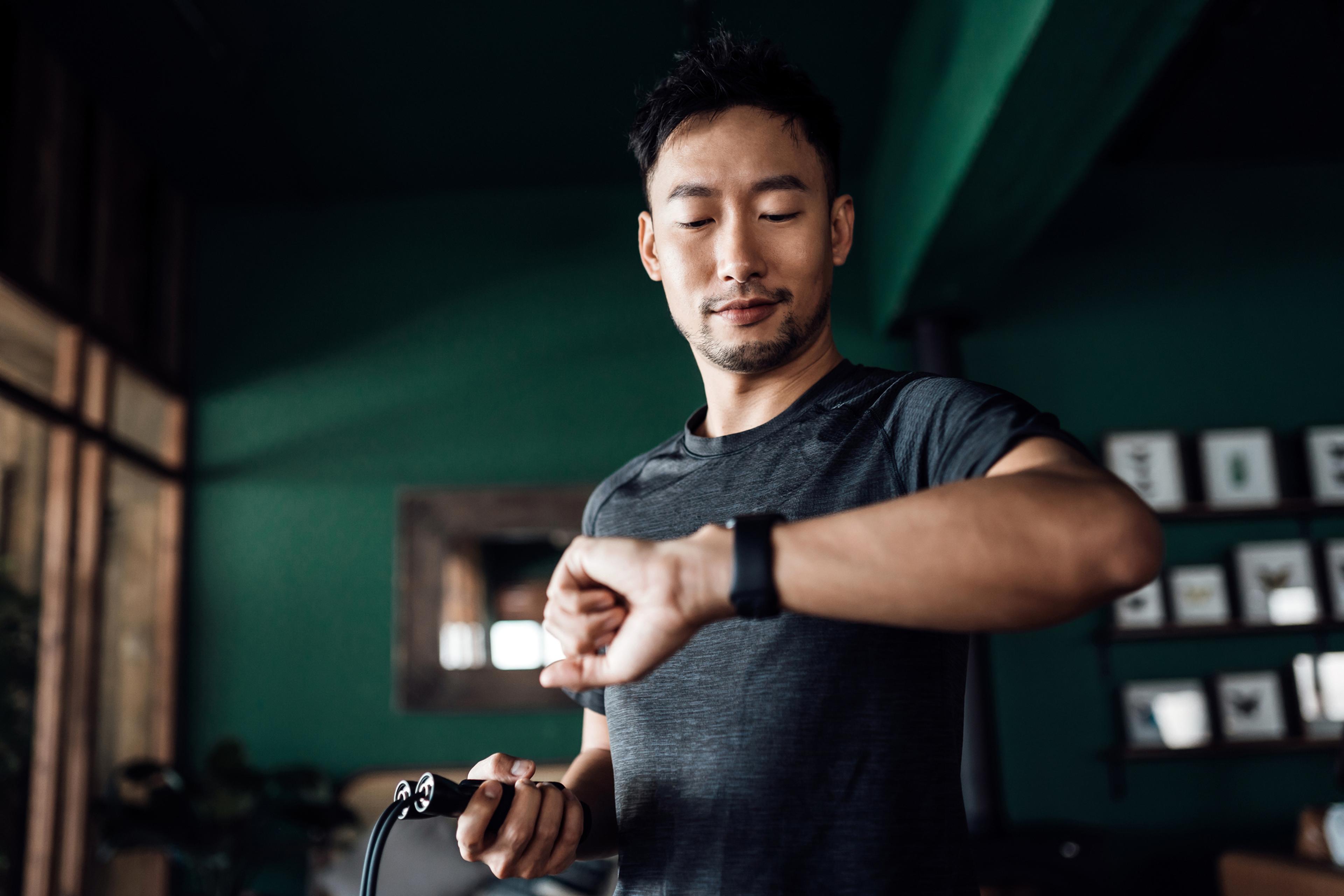 An active Asian man exercising at home uses a fitness tracker app on a smartwatch to monitor training progress and measure pulse.