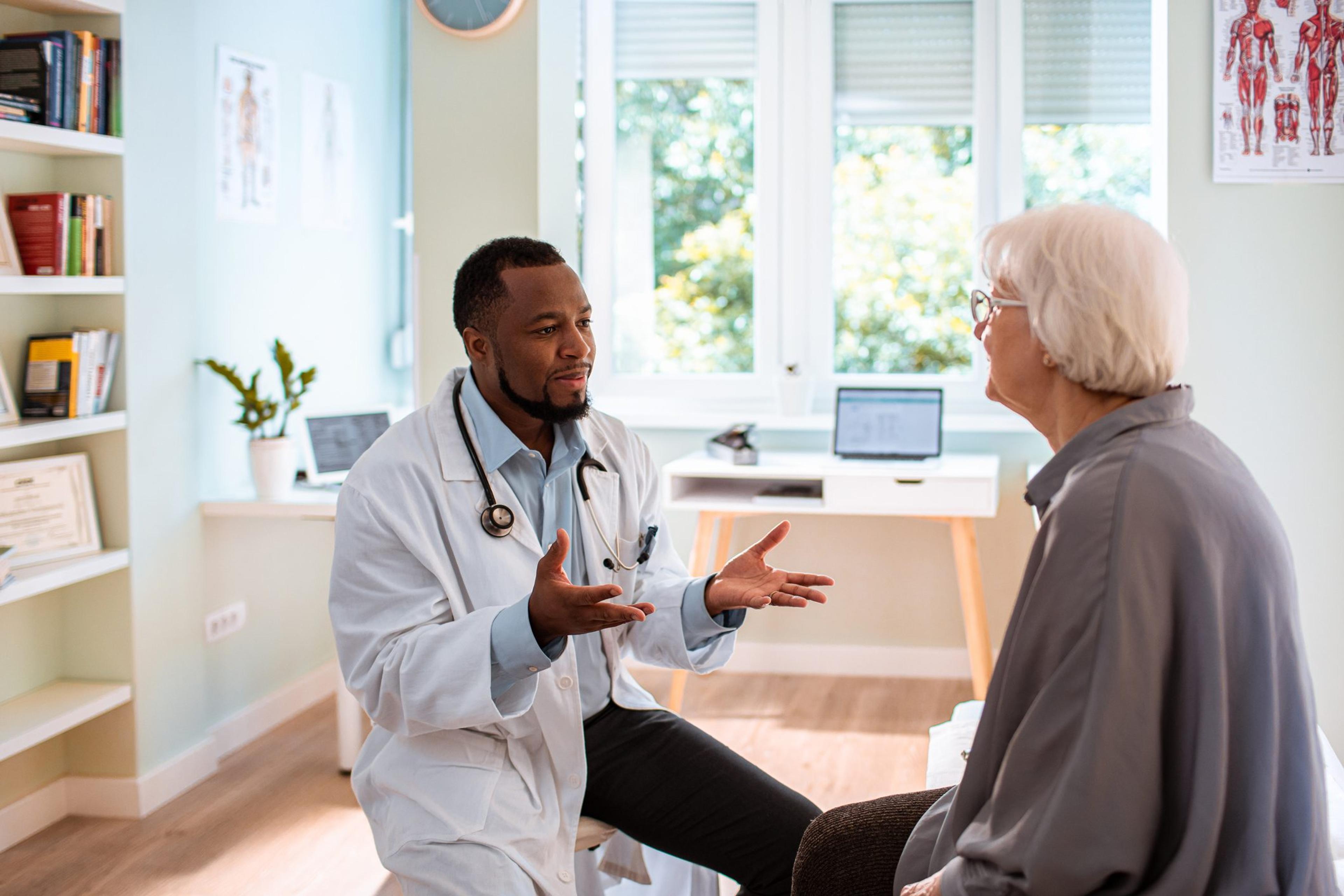 A young, Black, male doctor speaks with a white, elderly woman in a patient room with large windows and a lot of natural light.