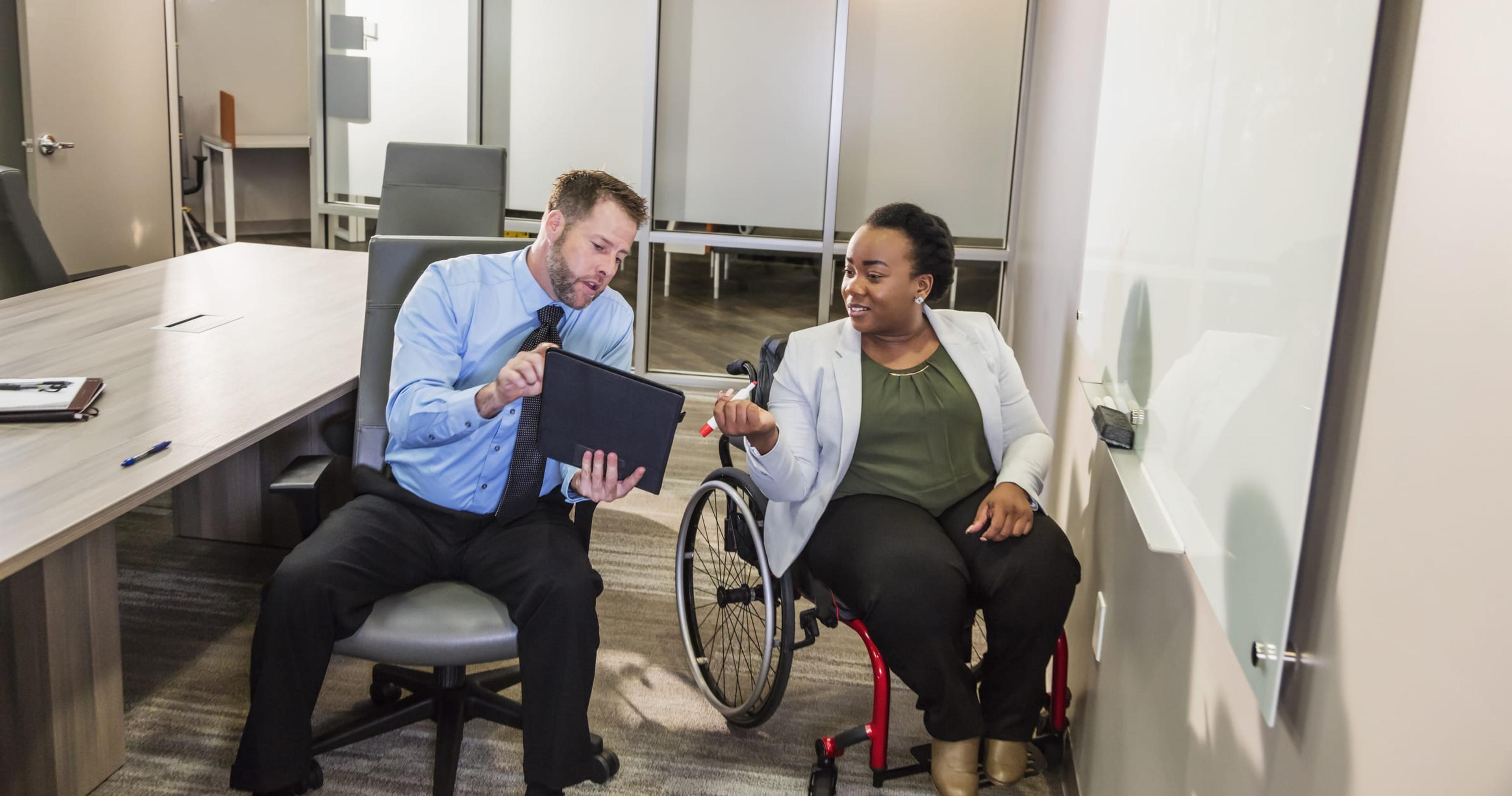 Two multi-ethnic business people in a meeting in an office boardroom. An African-American woman in a wheelchair is writing on a white board. Her coworker, a businessman wearing a shirt and tie, is holding up his notes for her to see. Both workers are in their 30s.