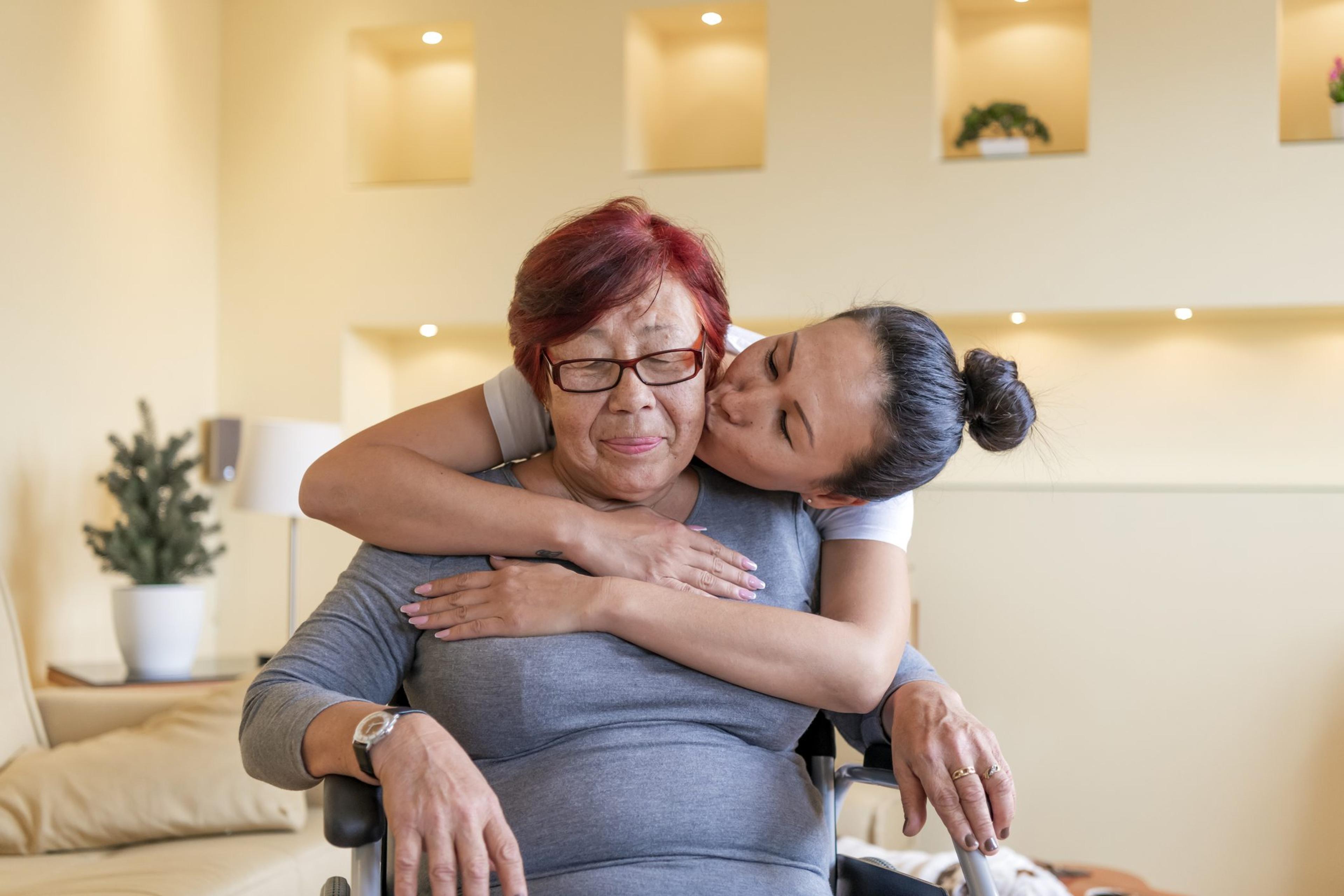 Woman with a disability sits in a chair and receives a kiss from her caregiver