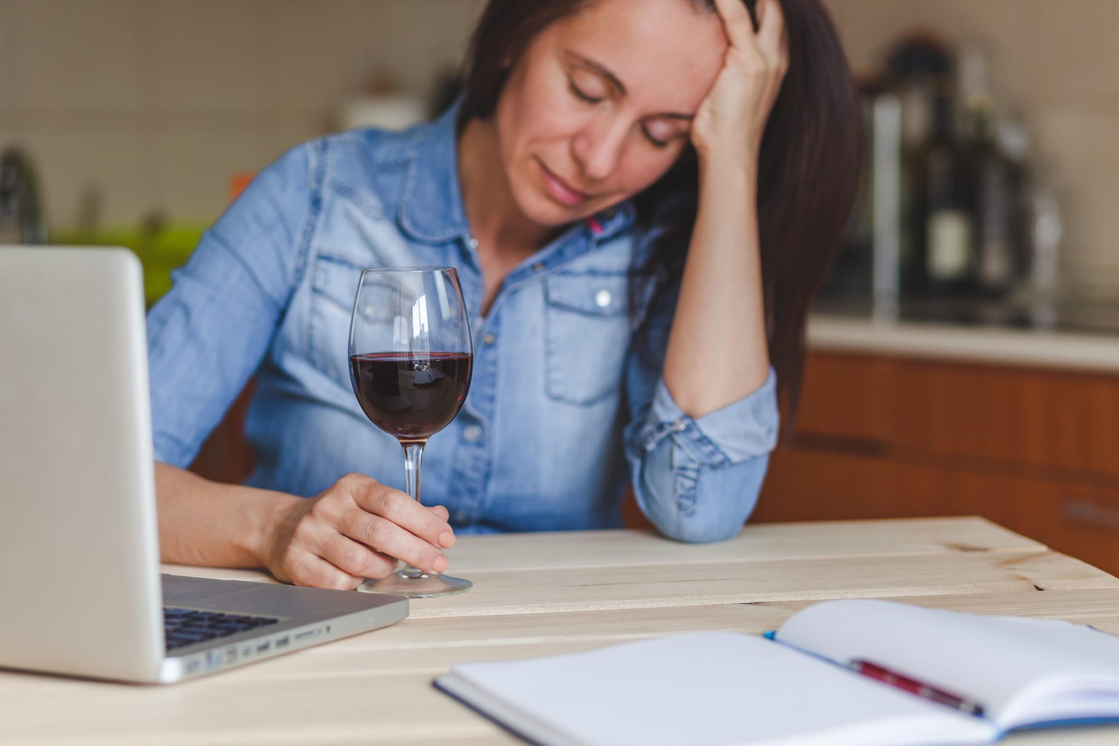 Overwhelmed, stressed woman sits with a glass of wine and her laptop