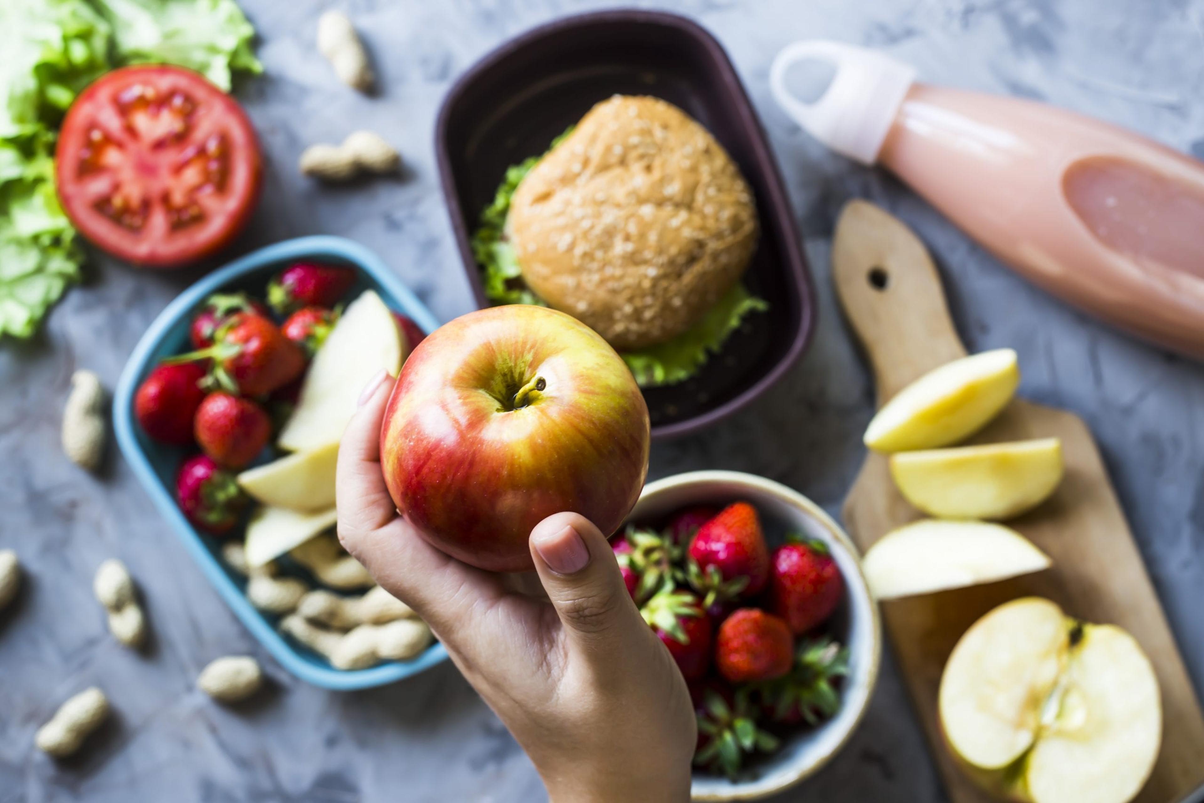 Cooking lunch for the child to school. On the gray kitchen table. Sandwich, strawberries and peanuts in lunchboxes. Top view. Woman holding an apple