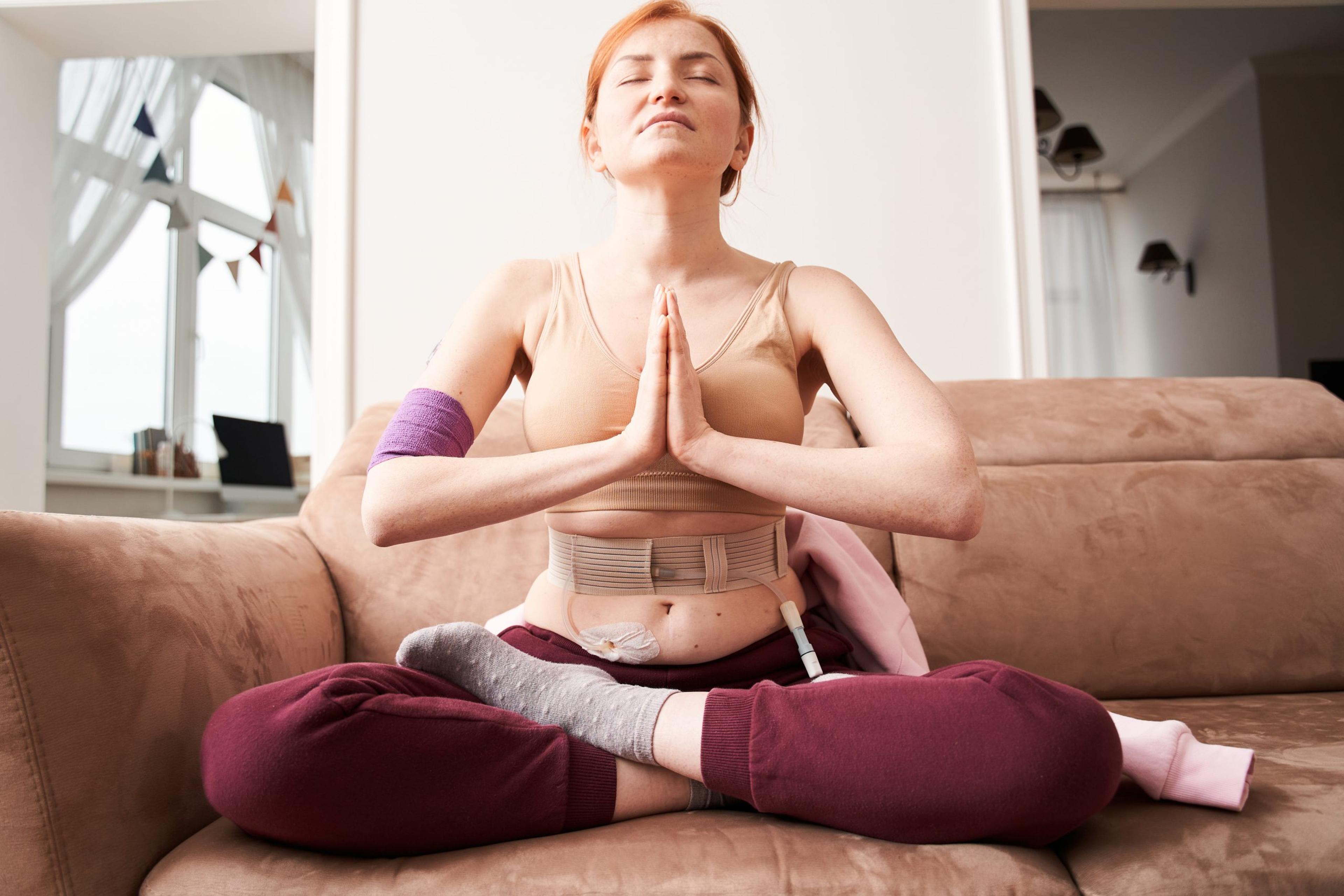 Full length mindful young woman with renal failure making praying gesture while sitting in lotus position at the sofa at home. Peaceful millennial girl deeply meditating, doing breathing yoga exercises alone