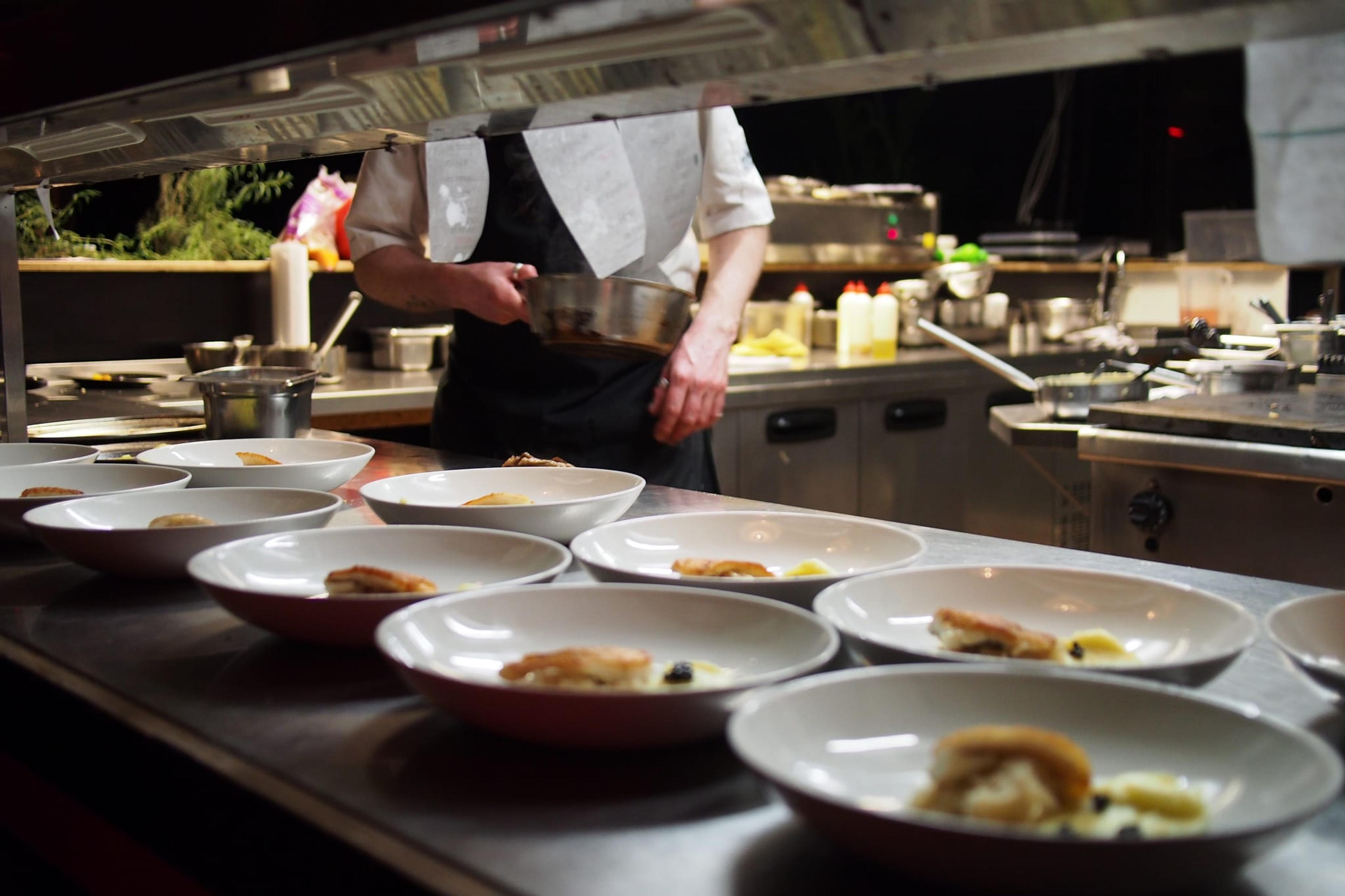 Photo of plated food in a restaurant kitchen, with chef in the background tossing food in a pan.