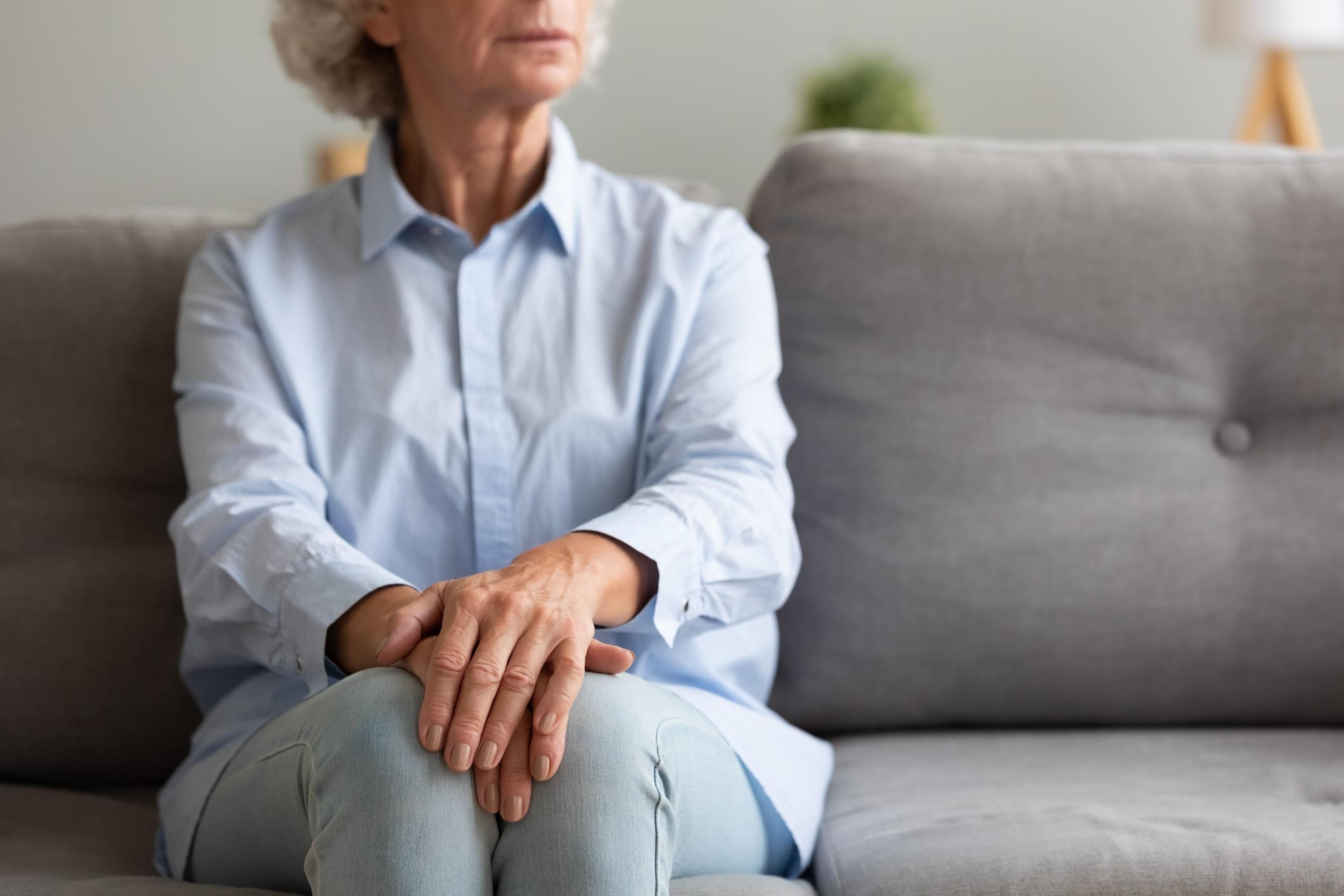 Unhappy depressed old senior woman sit alone on sofa with hands folded thinking of loneliness worried of disease or retirement problems feel anxiety grief suffer from arthritis concept, close up view