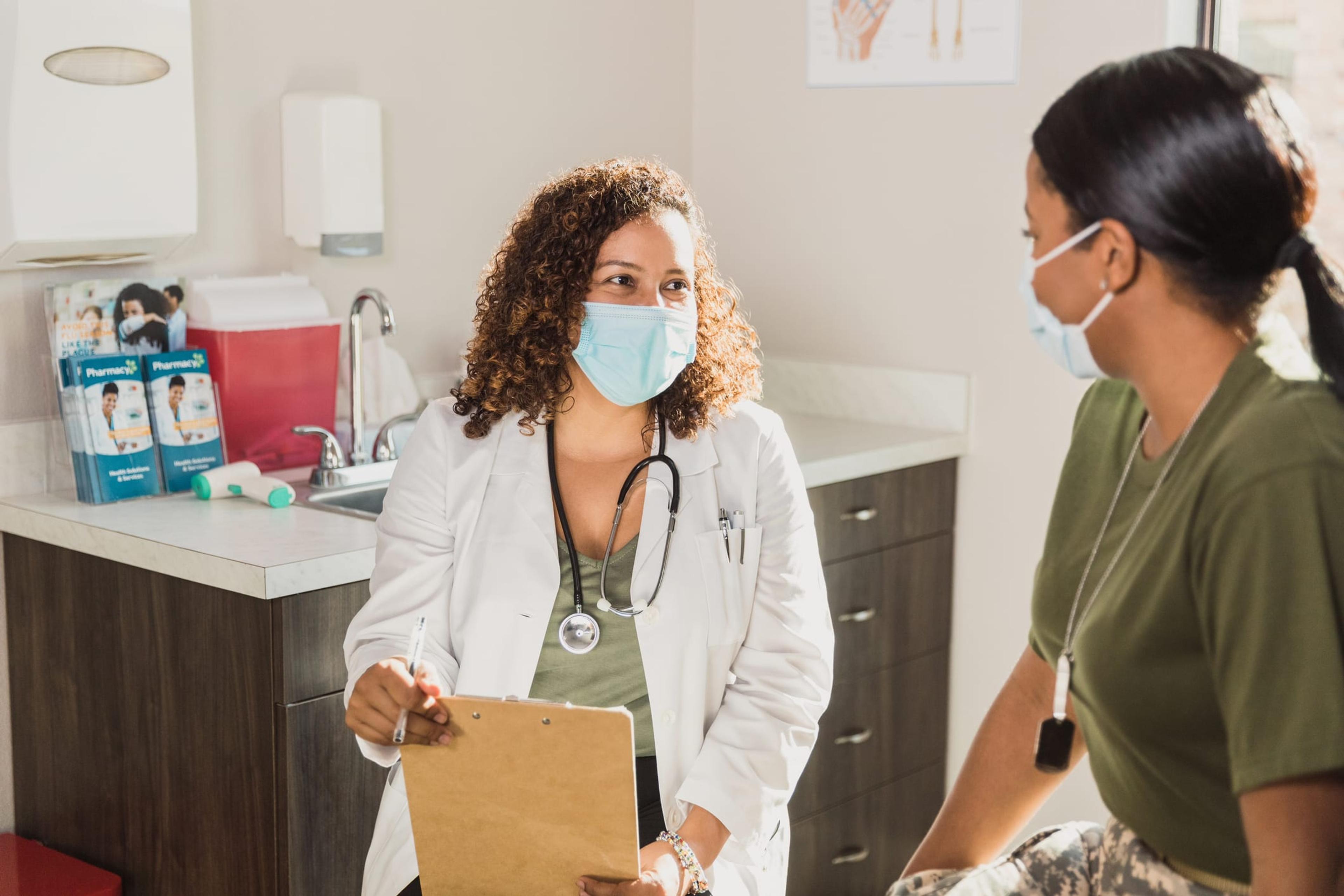 Woman in mask talking with a doctor holding a clipboard in a mask.