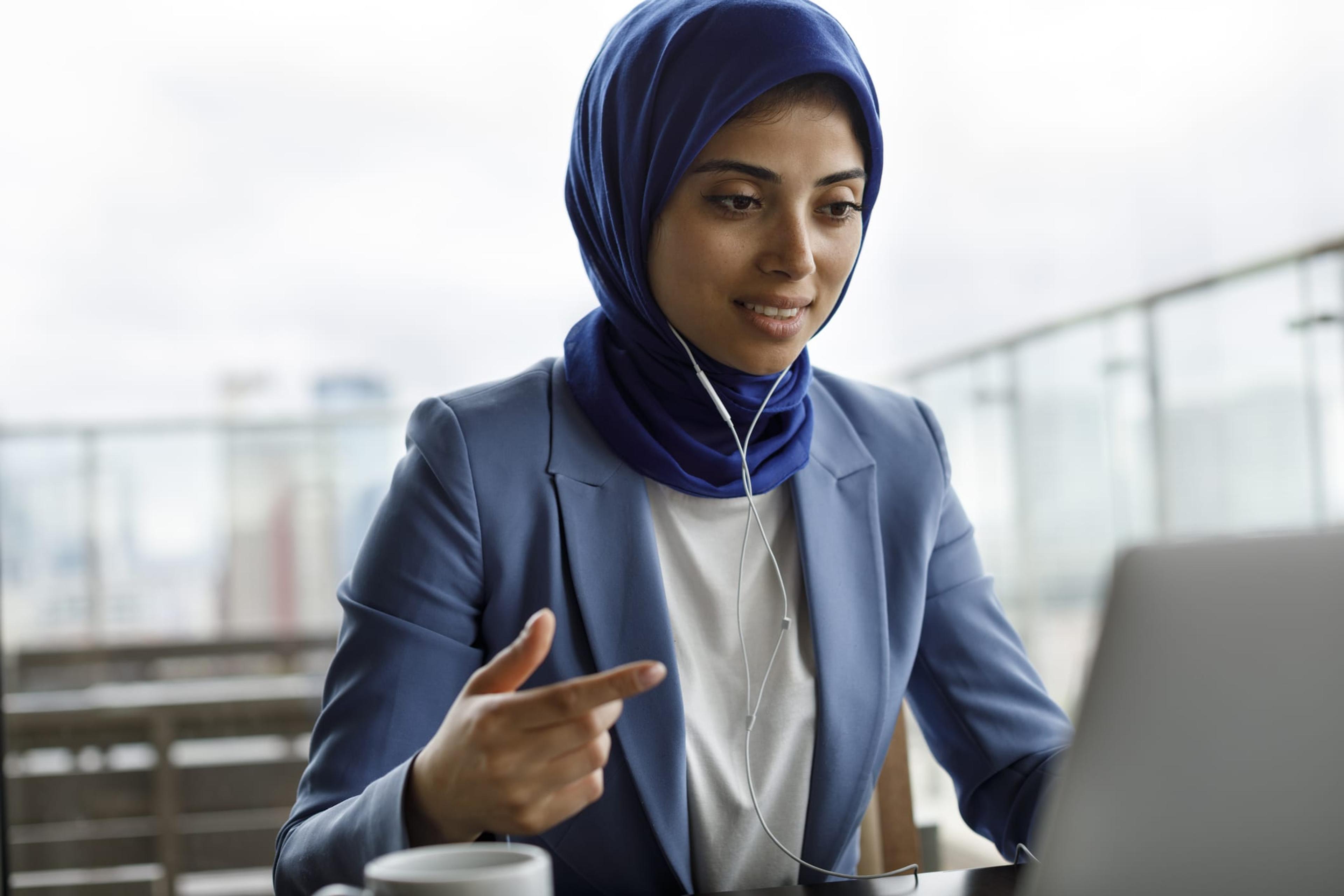 Young woman in blue suit and head covering motions to computer sitting in front of her.