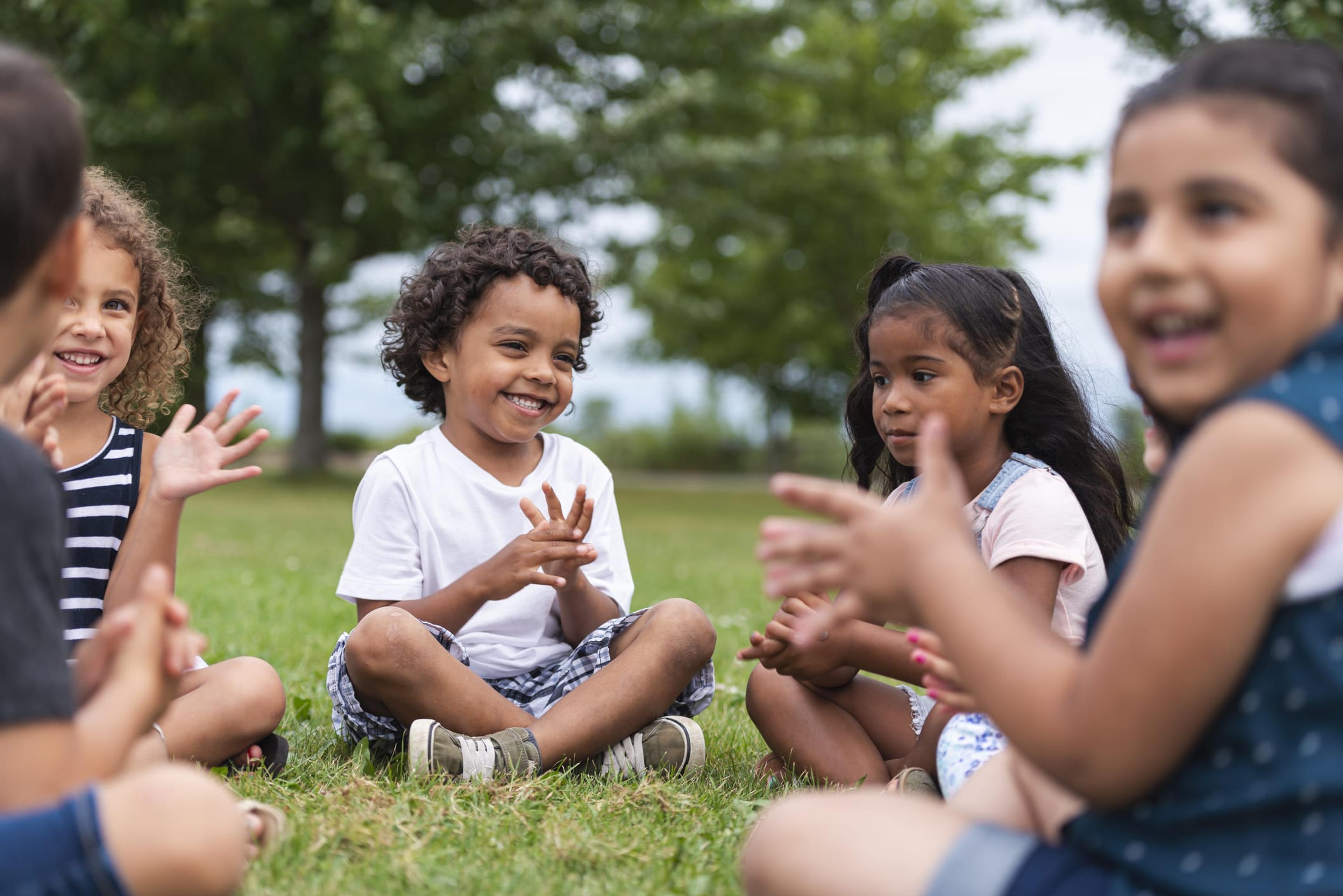 A group of multi-ethnic children sit in a circle outside and clap their hands