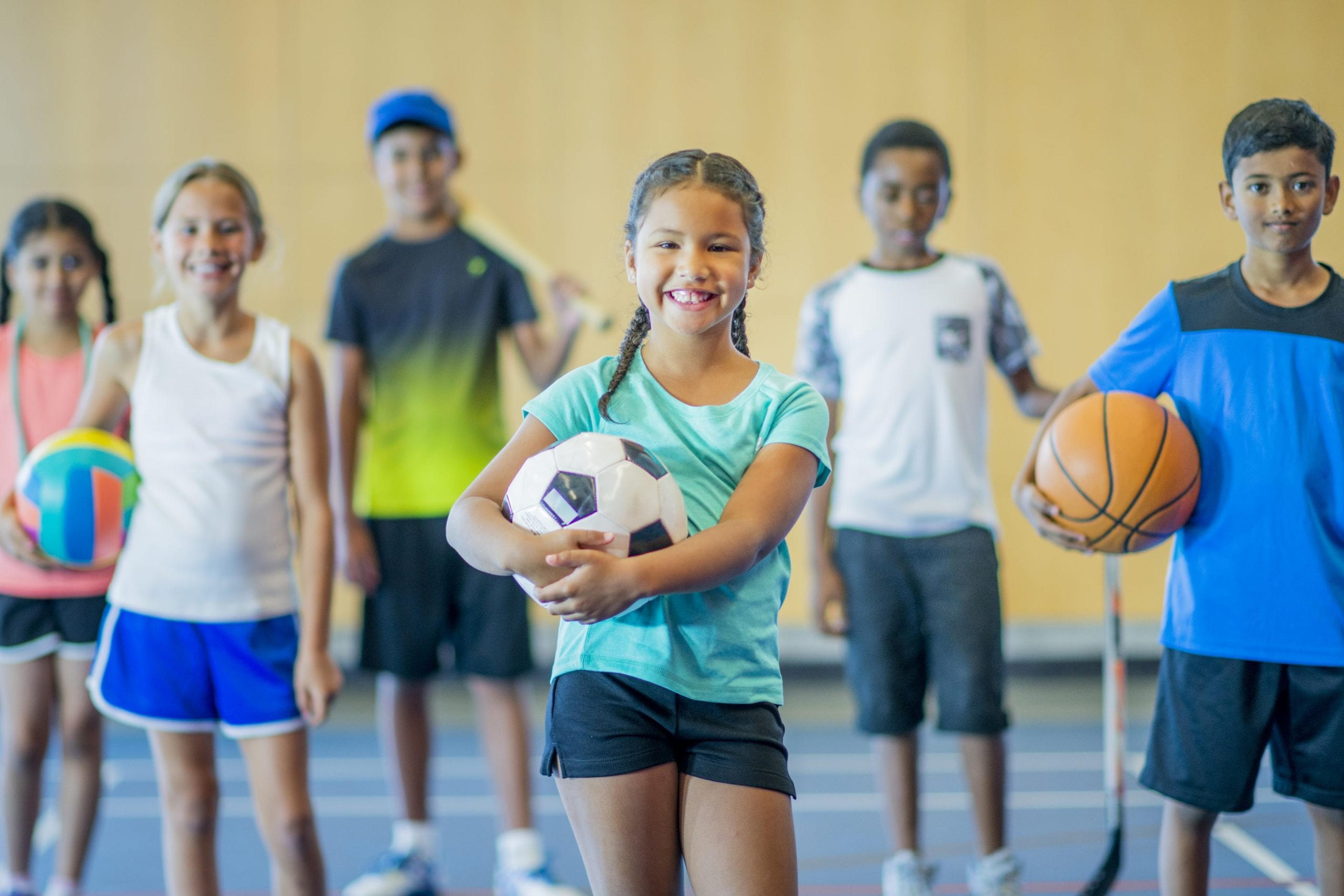 A multi-ethnic group of kids are standing in a gymnasium. They are holding a basketball, volleyball, hockey stick, skipping rope, baseball bat, and soccer ball. They are smiling.