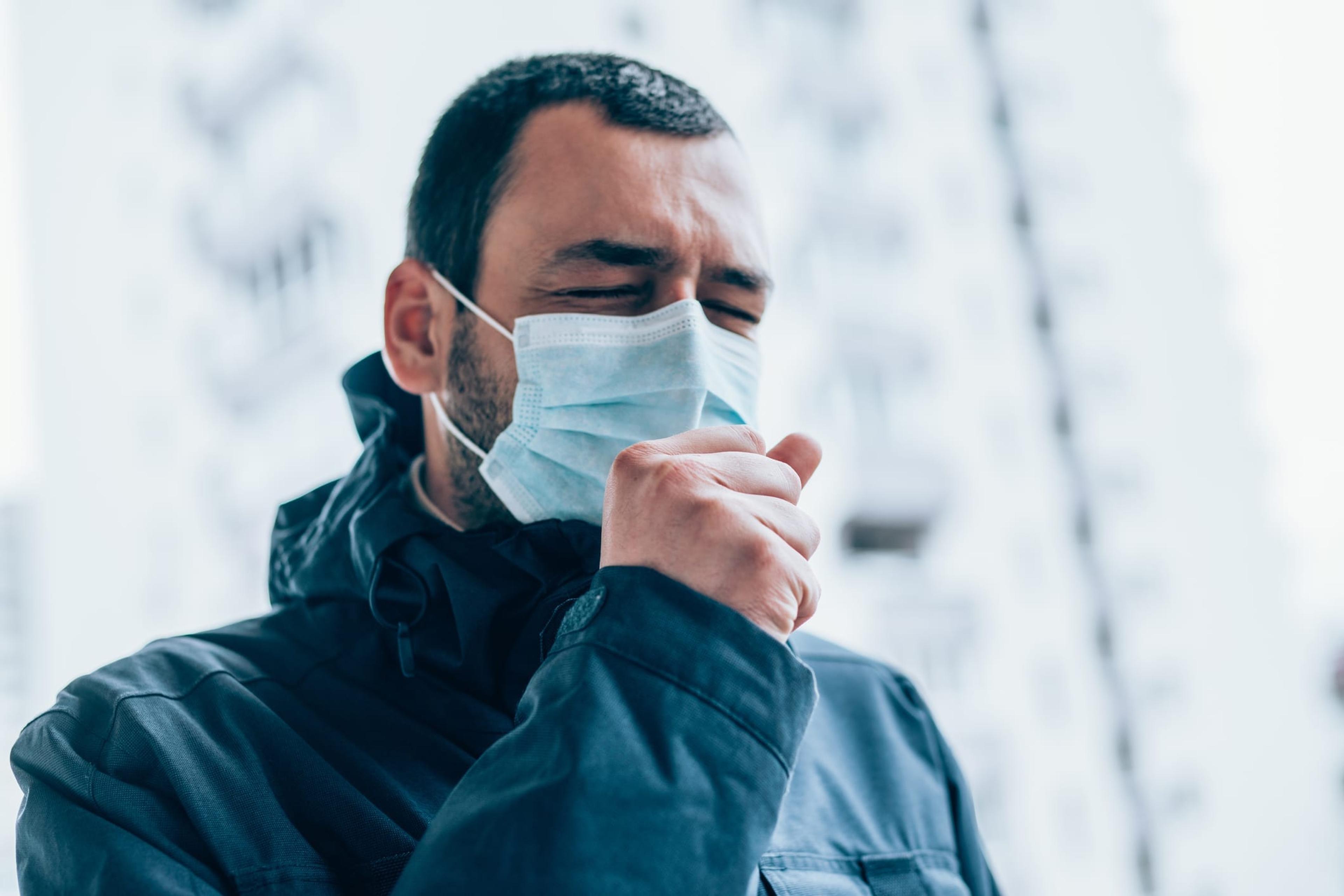 Portrait of young man on the street wearing face protective mask. Young man with closed eyes wear face protective mask and coughing in his arm.