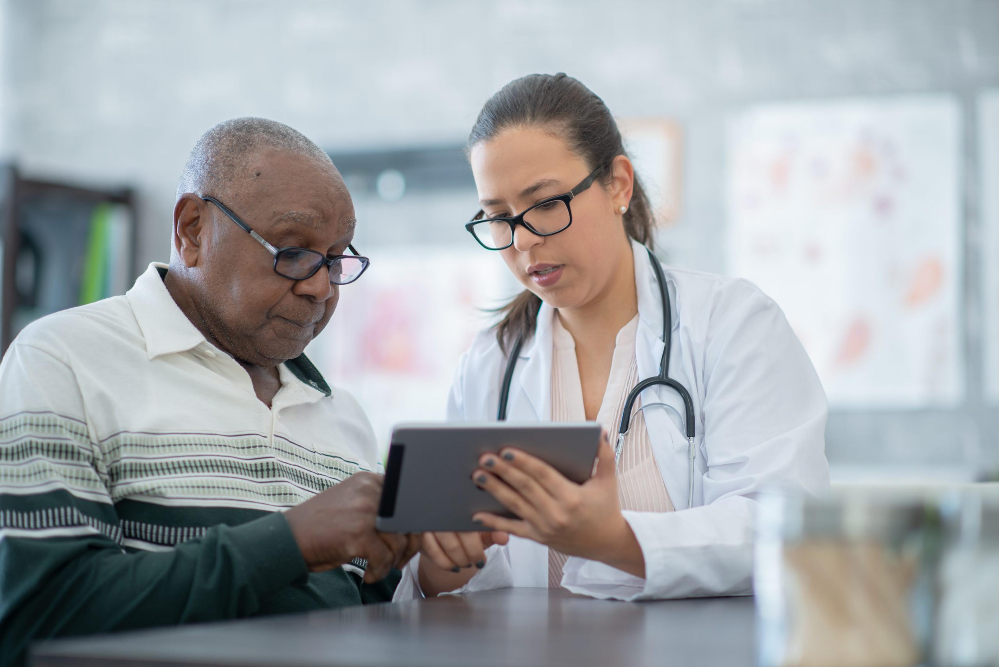 A Latina physician with a stethoscope around her neck shares a digital screen with a Black male patien