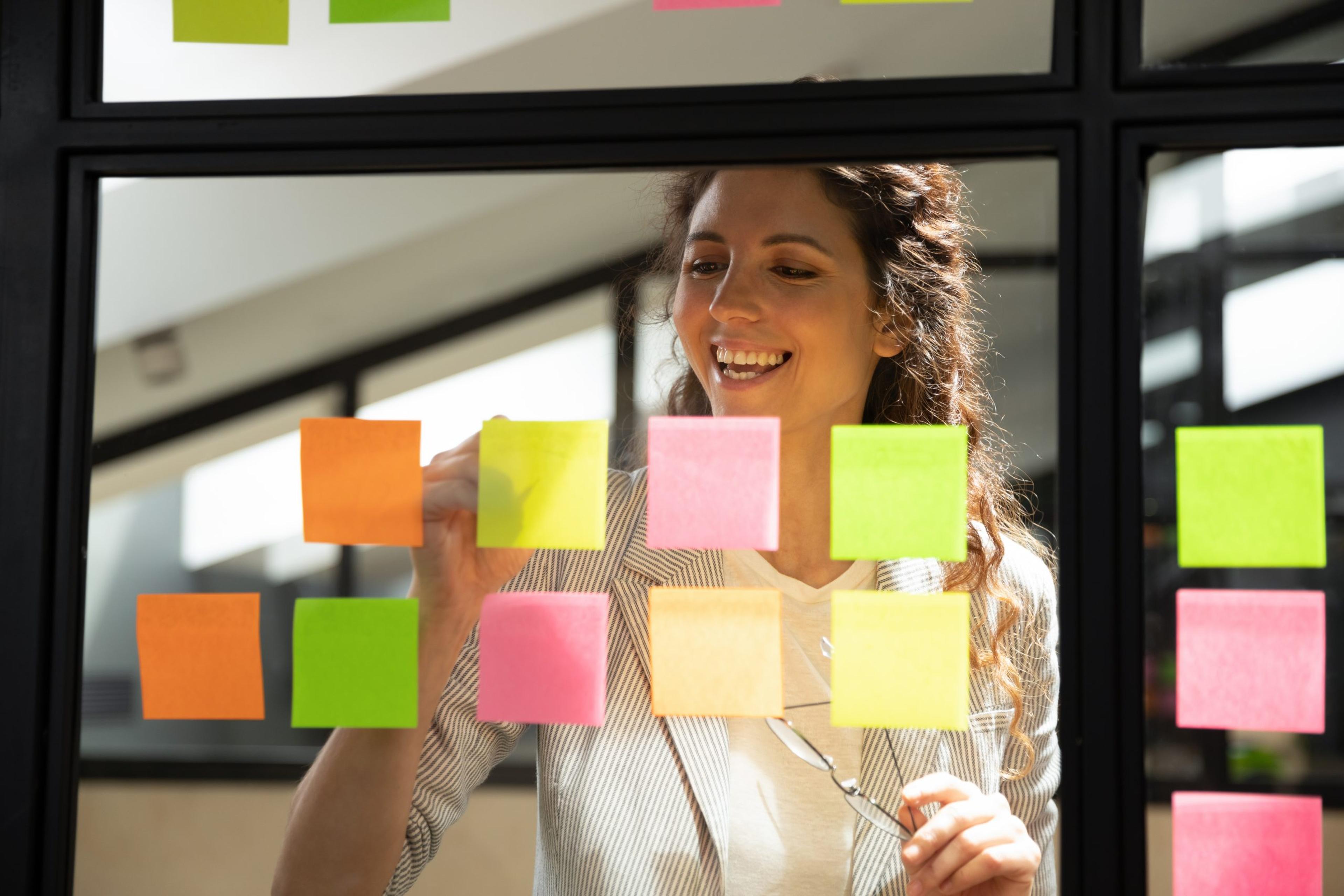 Attractive smiling businesswoman view through glass wall writing ideas creates personal tasks to-do list on post-it sticky notes, easy and effective way to manage and remember important issues concept