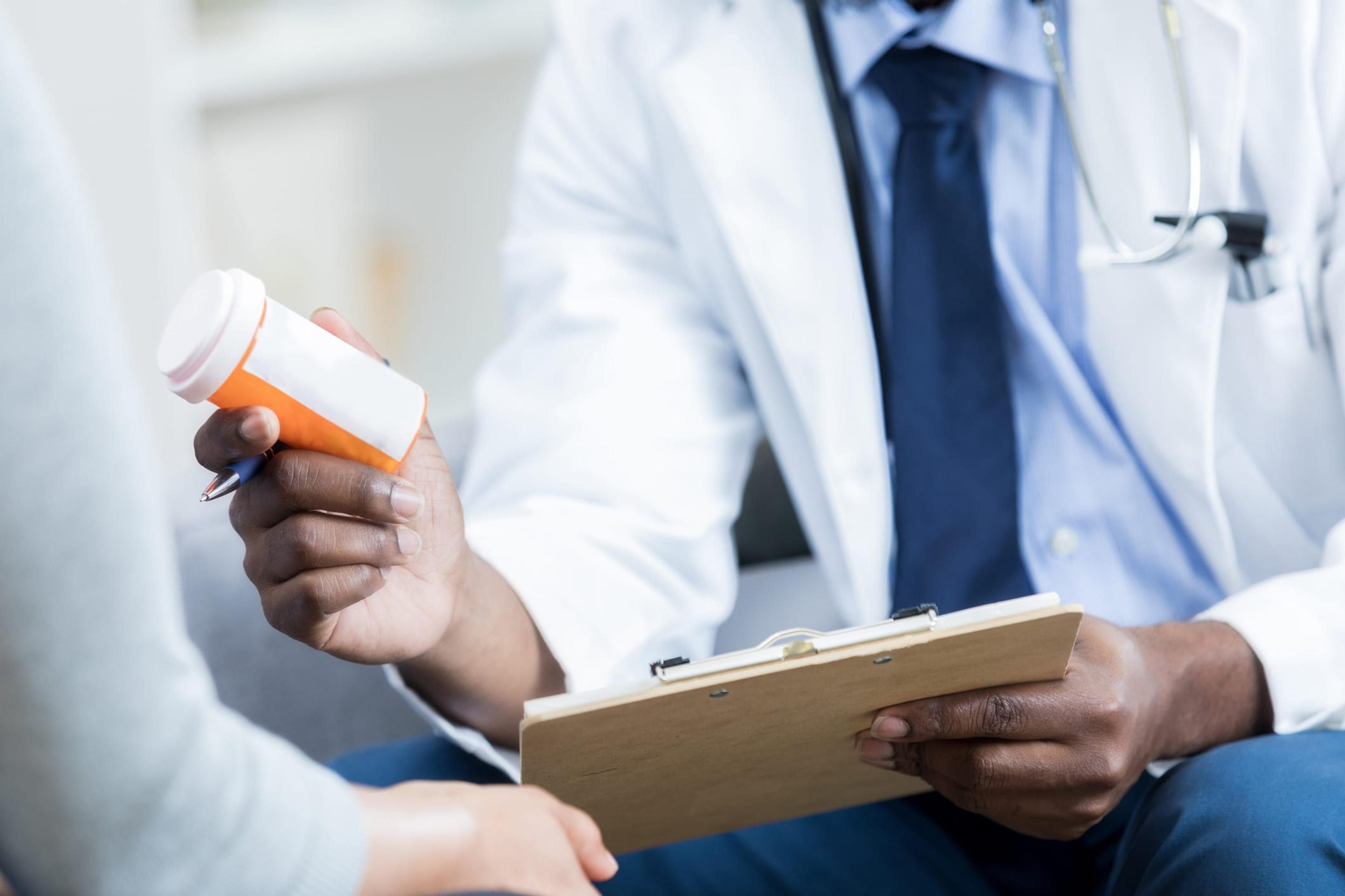 African American doctor hands a prescription medication bottle to a female patient. The doctor is holding the patient's chart.