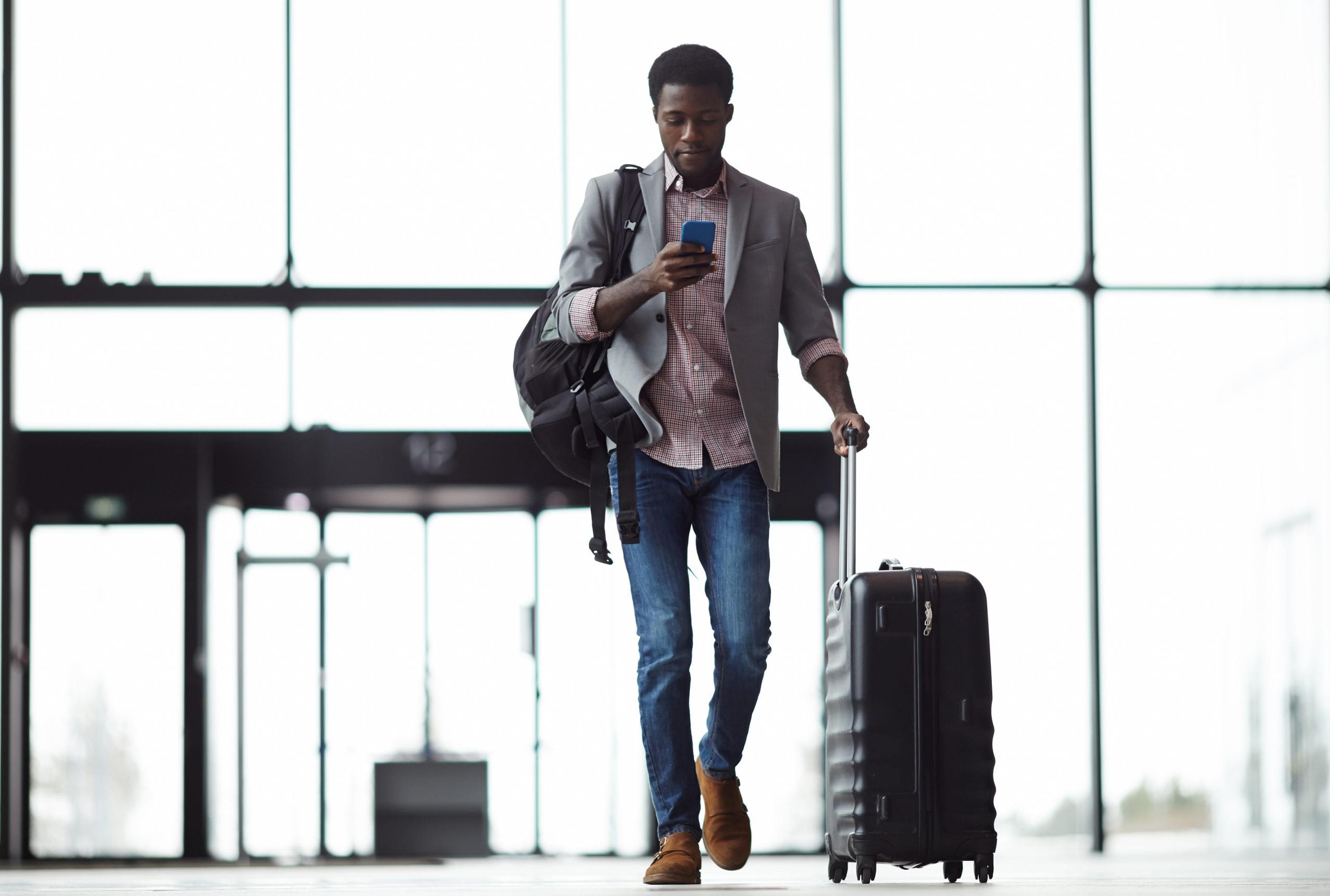 Young man checking his phone as he wheels a suitcase through the airport.