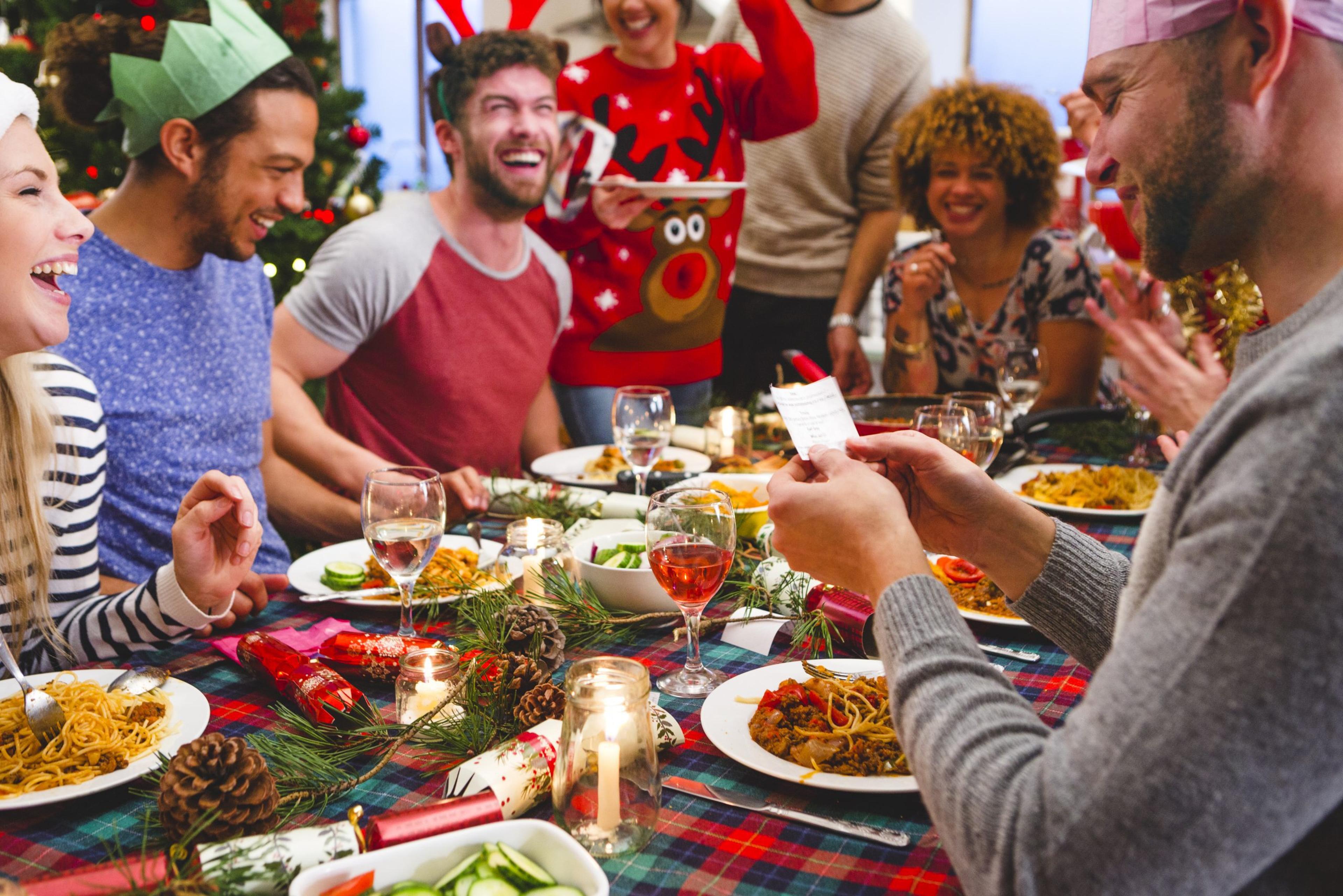 Group of friends laughing as they sit around a table eating and drinking at Christmas. One man is reading a joke from a christmas cracker. Party hats and christmas jumpers are worn.