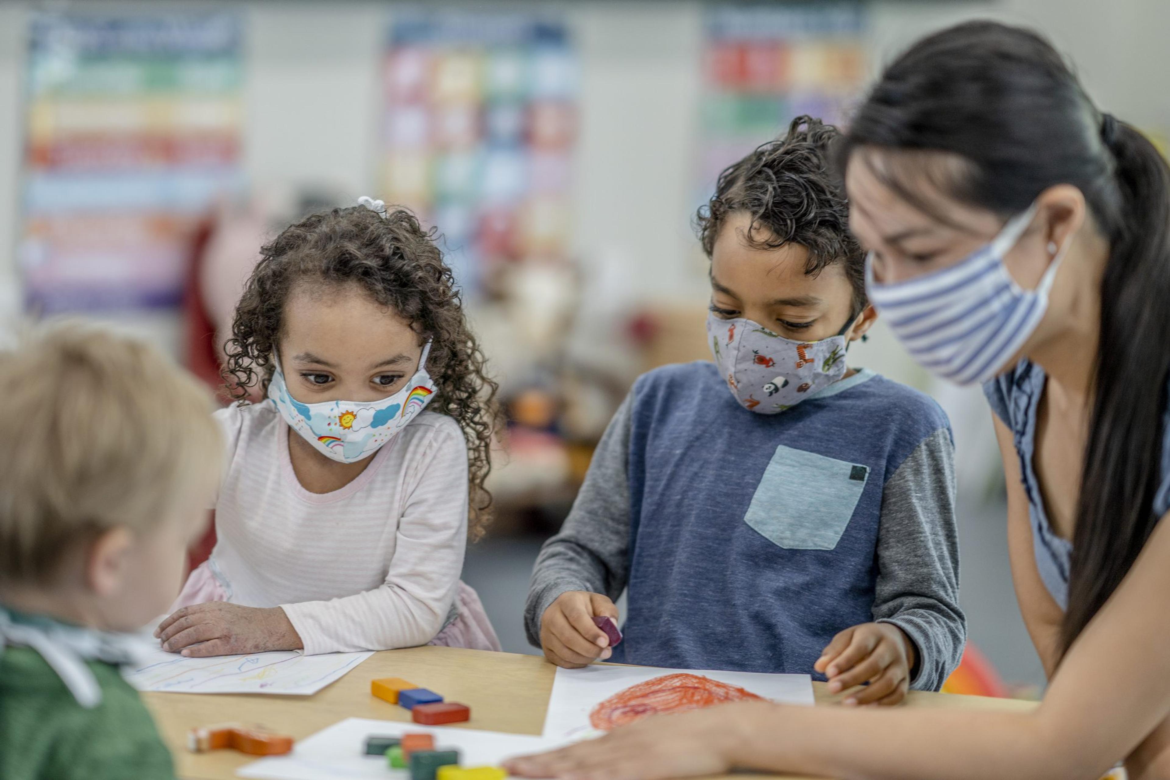 Three masked children sit around a table in a school setting with blocks and a masked female teacher leading them.