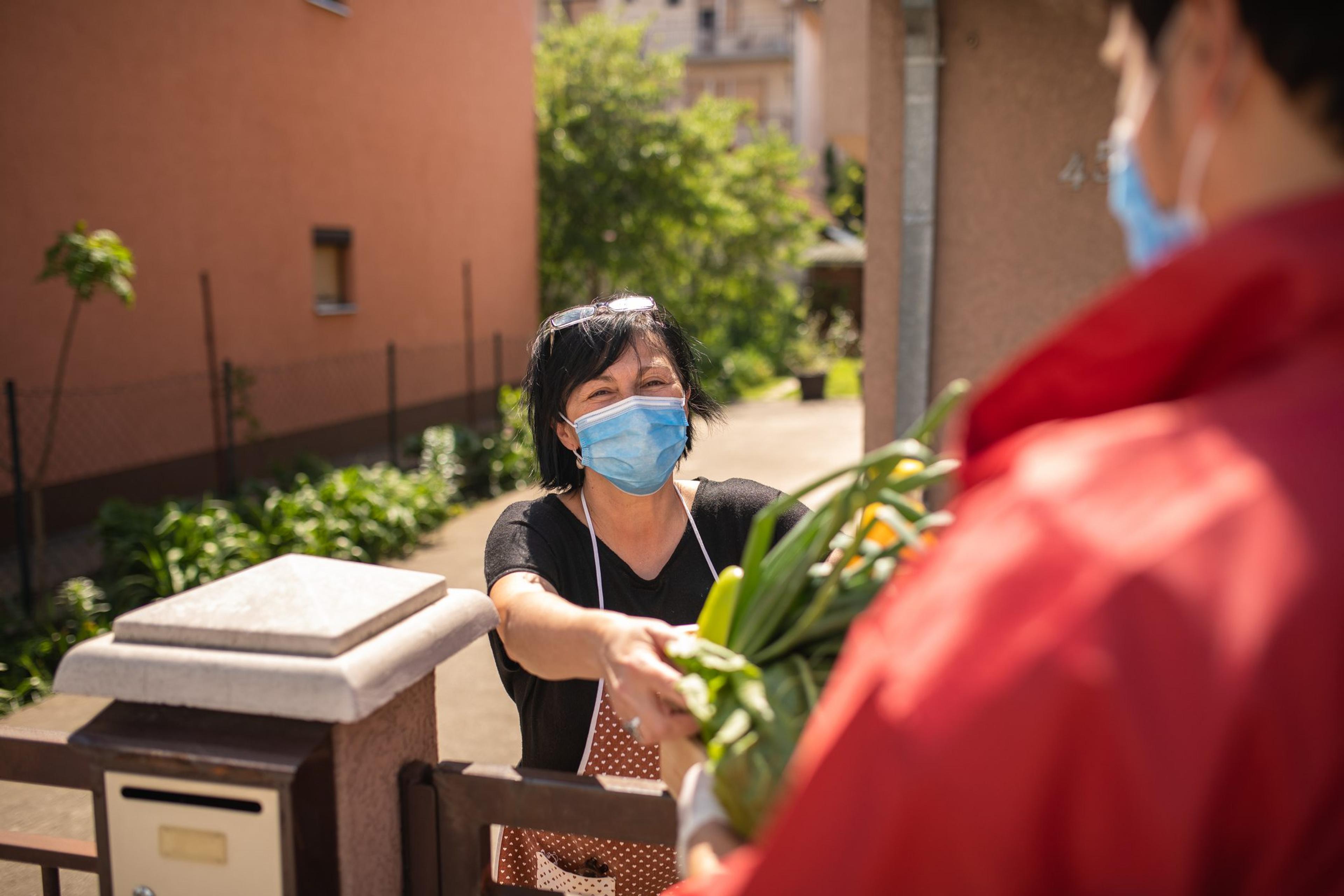 Senior woman is grateful for food delivery from helpful volunteer during covid-19, they are wearing protective masks