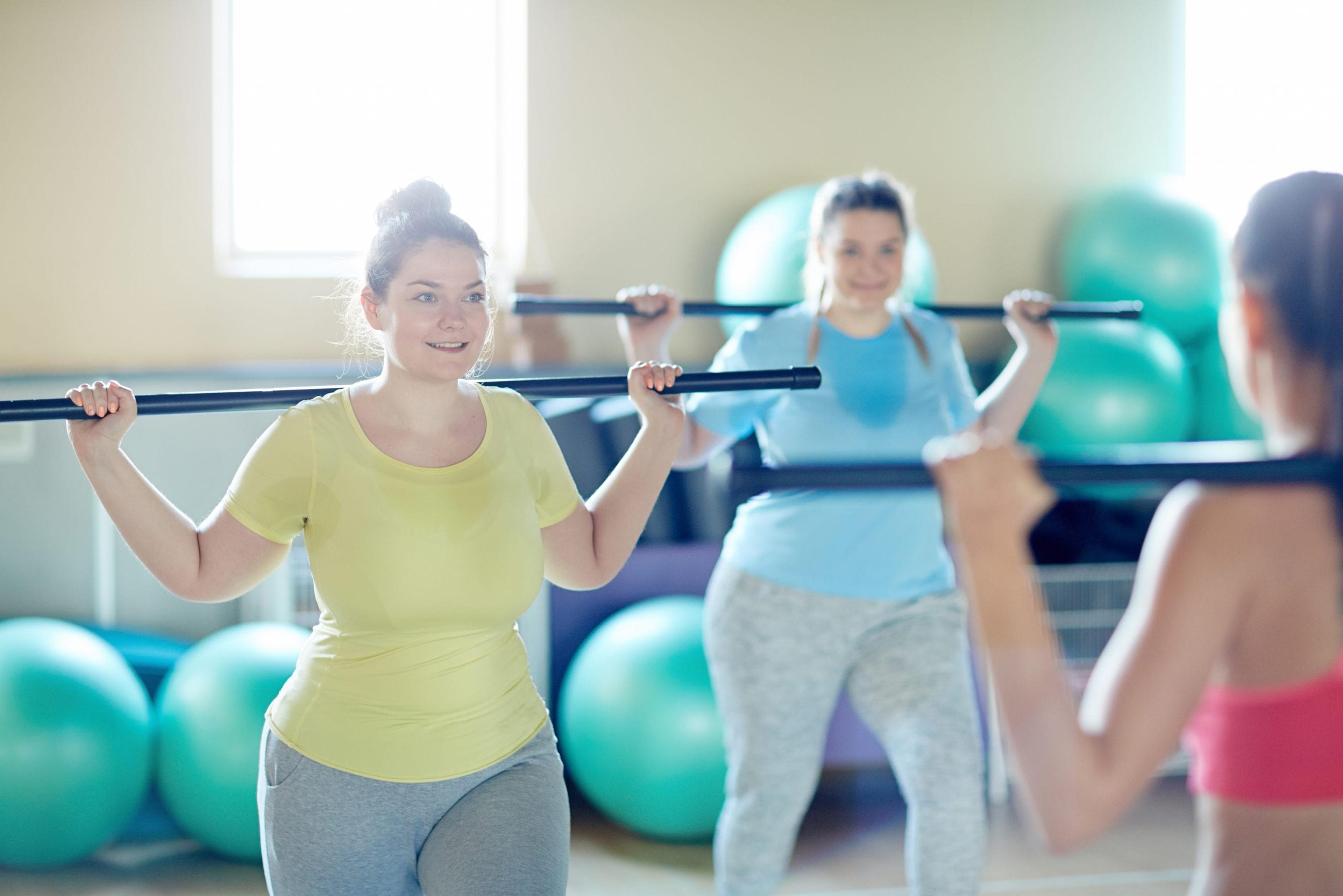 Two full figured women with gymnastic bars doing exercises after their instructor in gym