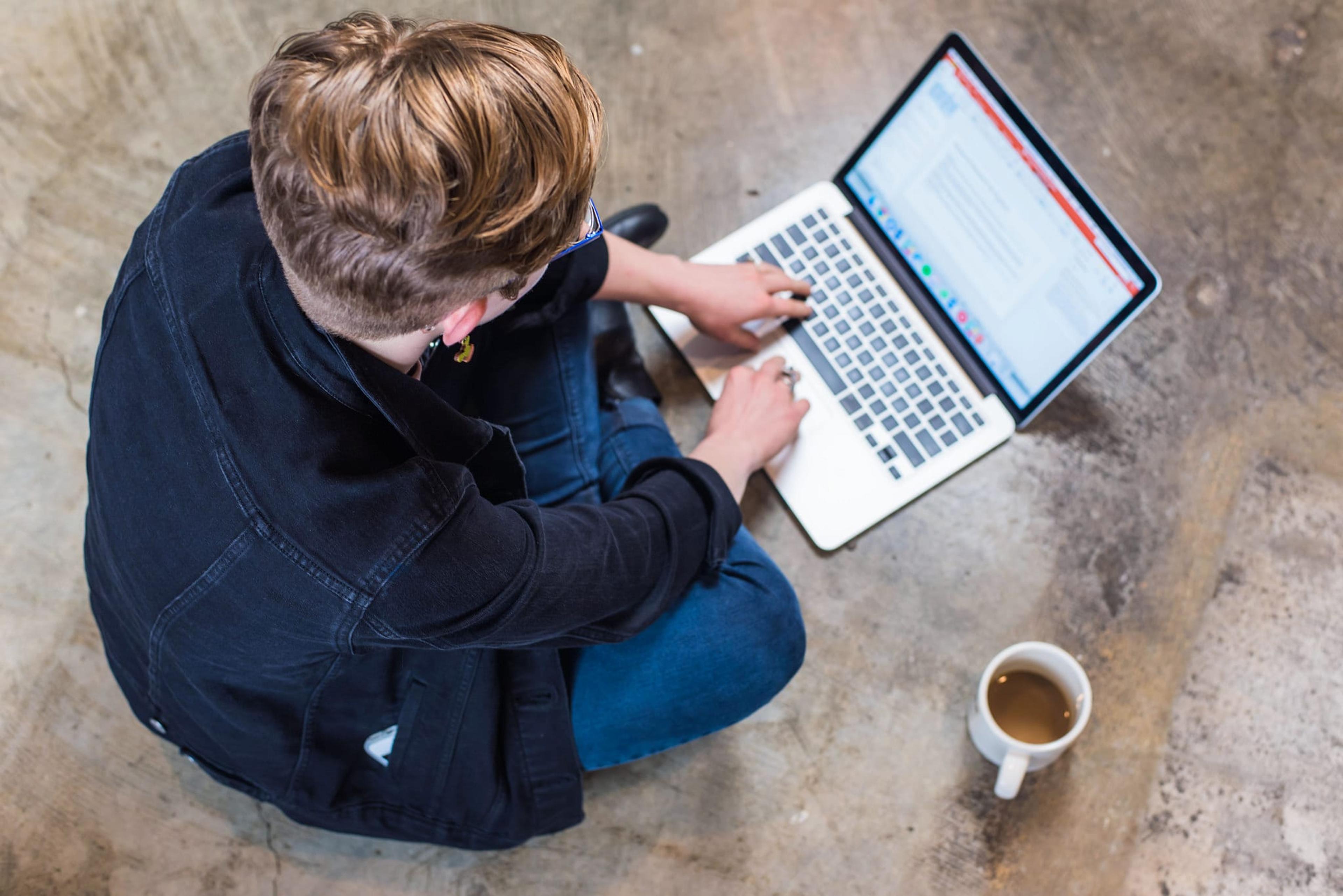 A woman sits cross-legged, typing on her laptop, which sits in front of her on the floor. A cup of coffee is on the ground to her side.