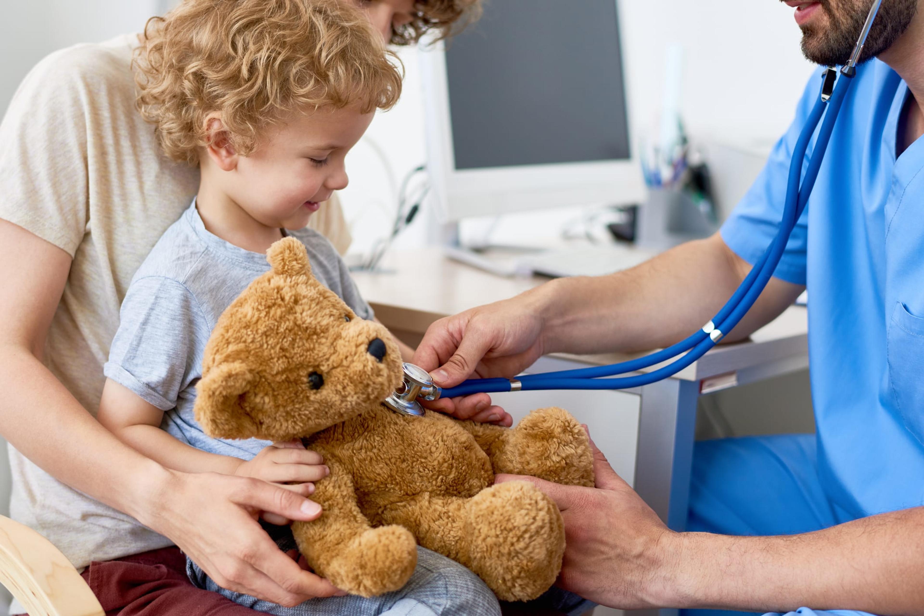 Image of doctor using stethoscope on teddy bear being held by toddler in his mother's lap.
